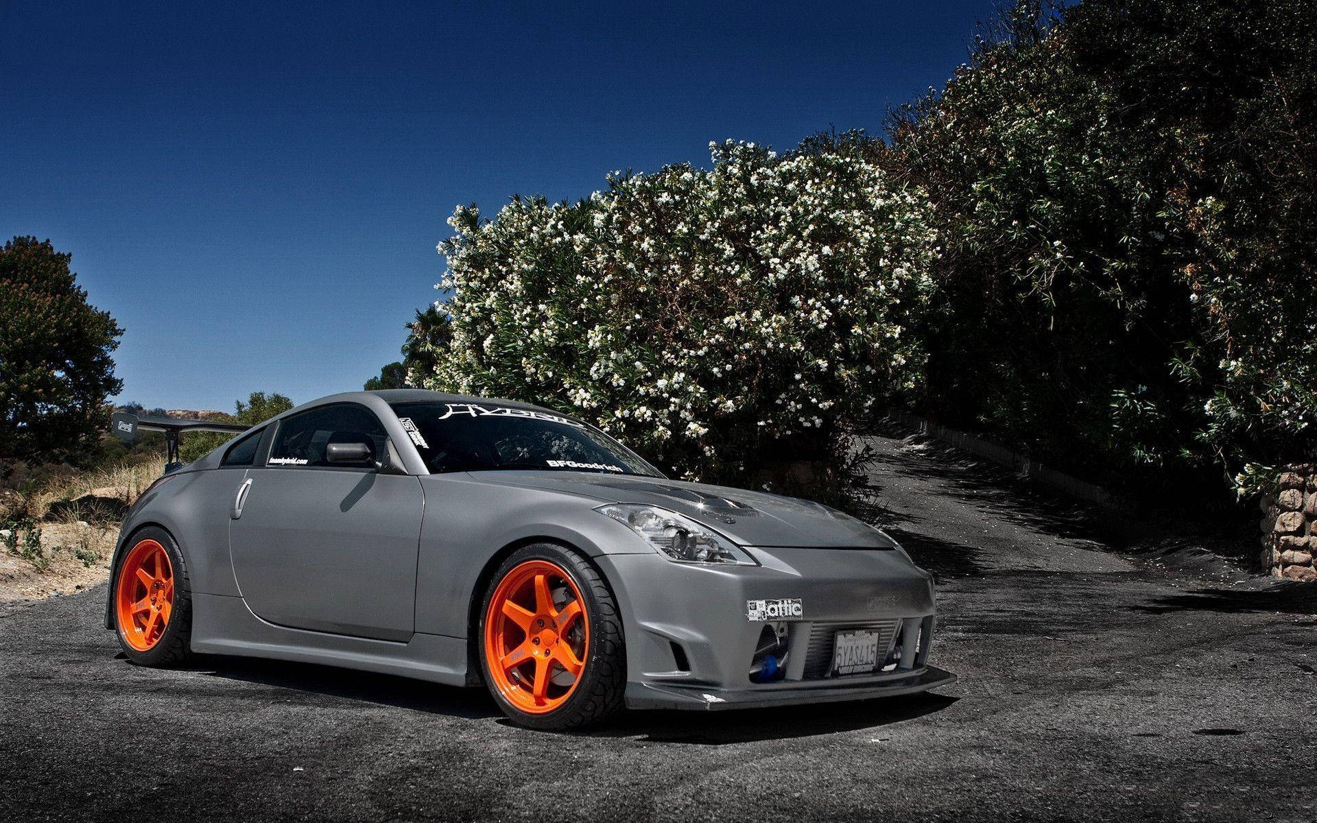 駕駛 Nissan 350Z 畅遊開放道路 壁紙 — 一輛啞光灰色跑車配以鮮豔橙色車輪，停泊在碎石車道上，旁邊是盛開的白色灌木叢，上方是清澈的藍天。