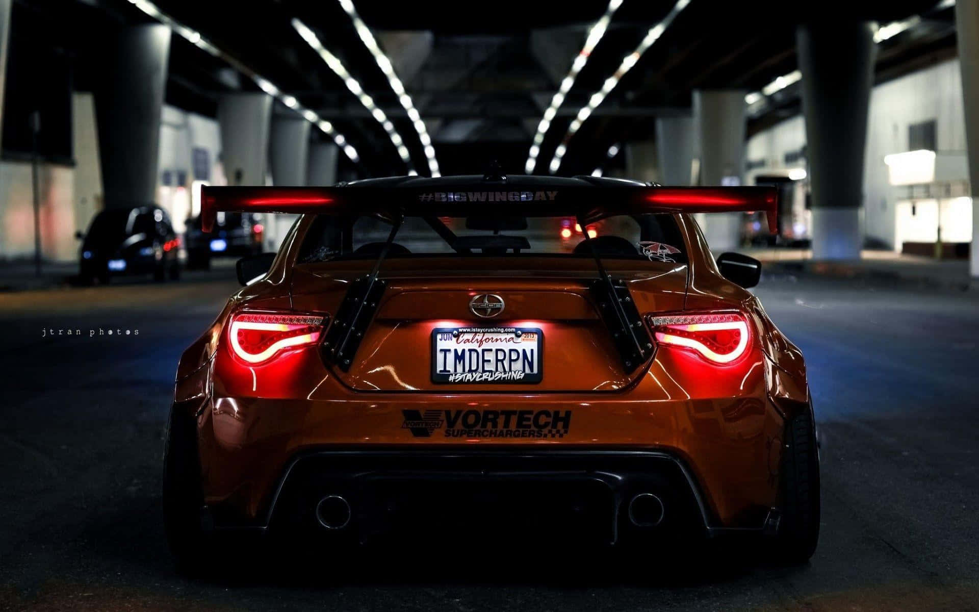 駕駛 Toyota 86，突破極限 壁紙 — 一輛定製的橙色跑車，帶有發光的紅色尾燈，夜間停放在光線昏暗的城市天橋下。