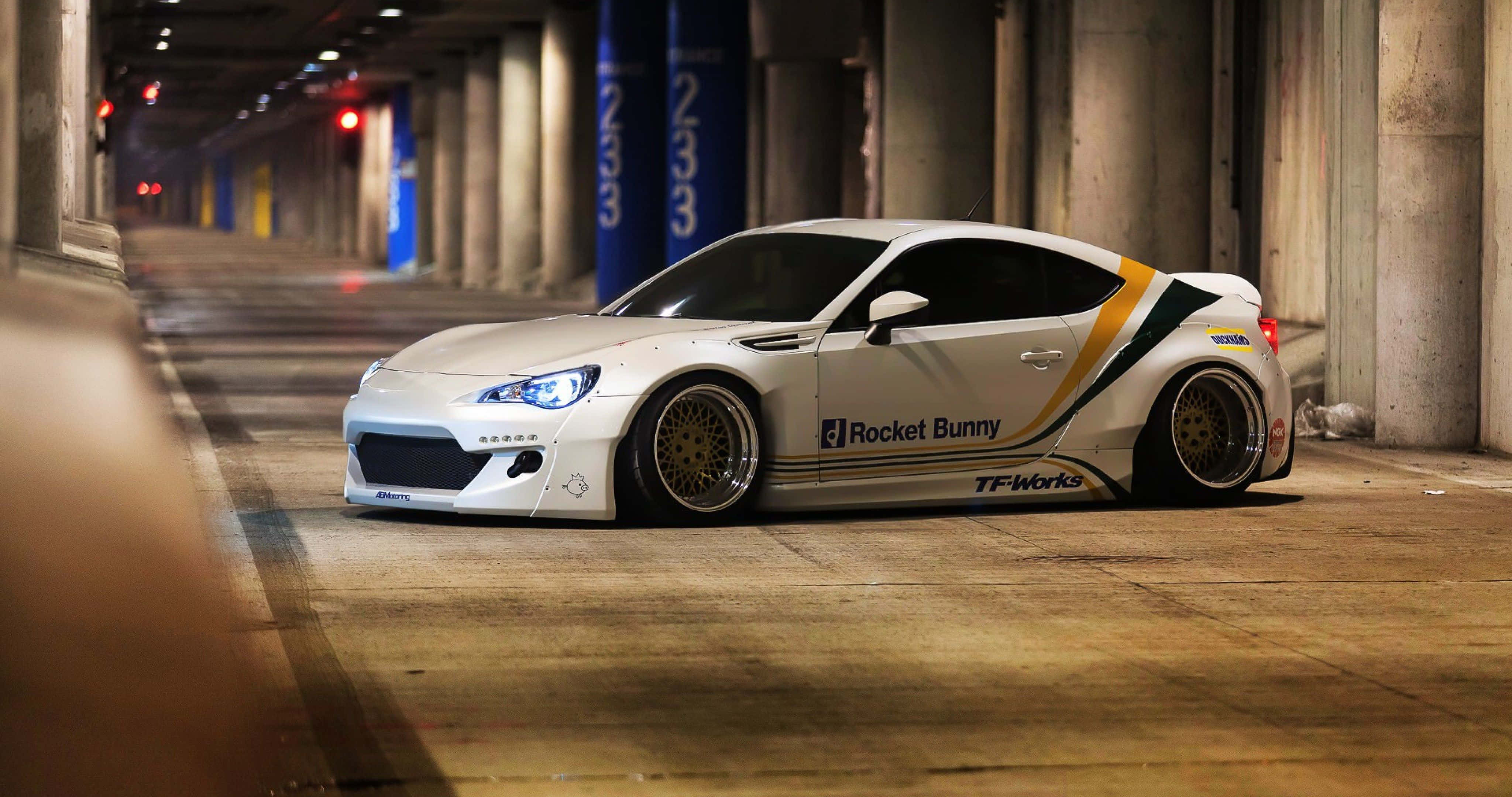 駕駛 Toyota 86，輕鬆過彎 壁紙 — 夜晚，一輛白色Rocket Bunny改裝跑車配金色輪圈停泊在光線昏暗的混凝土天橋下。