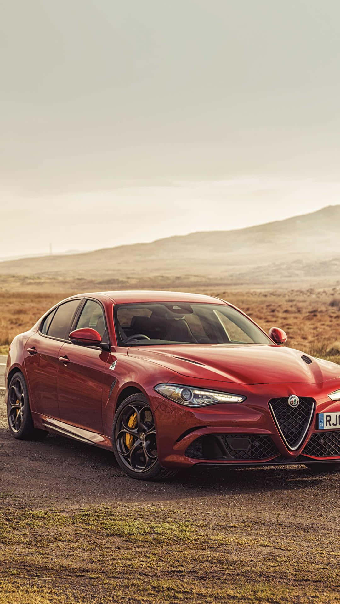 駕駛Alfa Romeo，享受奢華之旅 壁紙 — 一輛亮紅色的Alfa Romeo轎車停泊在塵土飛揚的道路上，霧濛濛的天空下是起伏的丘陵。