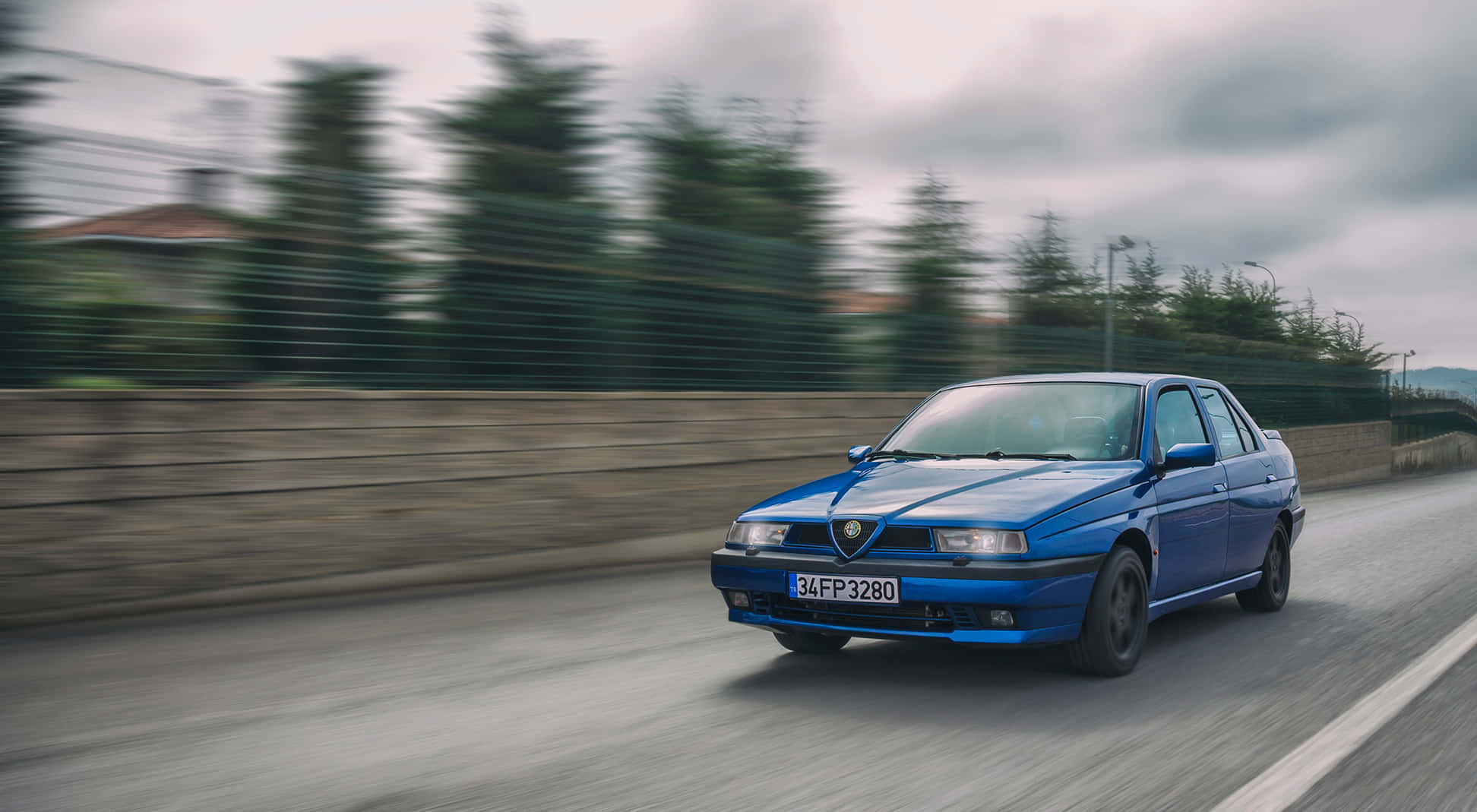 駕駛Alfa Romeo，穿梭城市街頭 壁紙 — 一輛充滿活力的藍色愛快羅密歐145在路邊疾馳，帶有動態模糊，在陰天傳達動態運動。