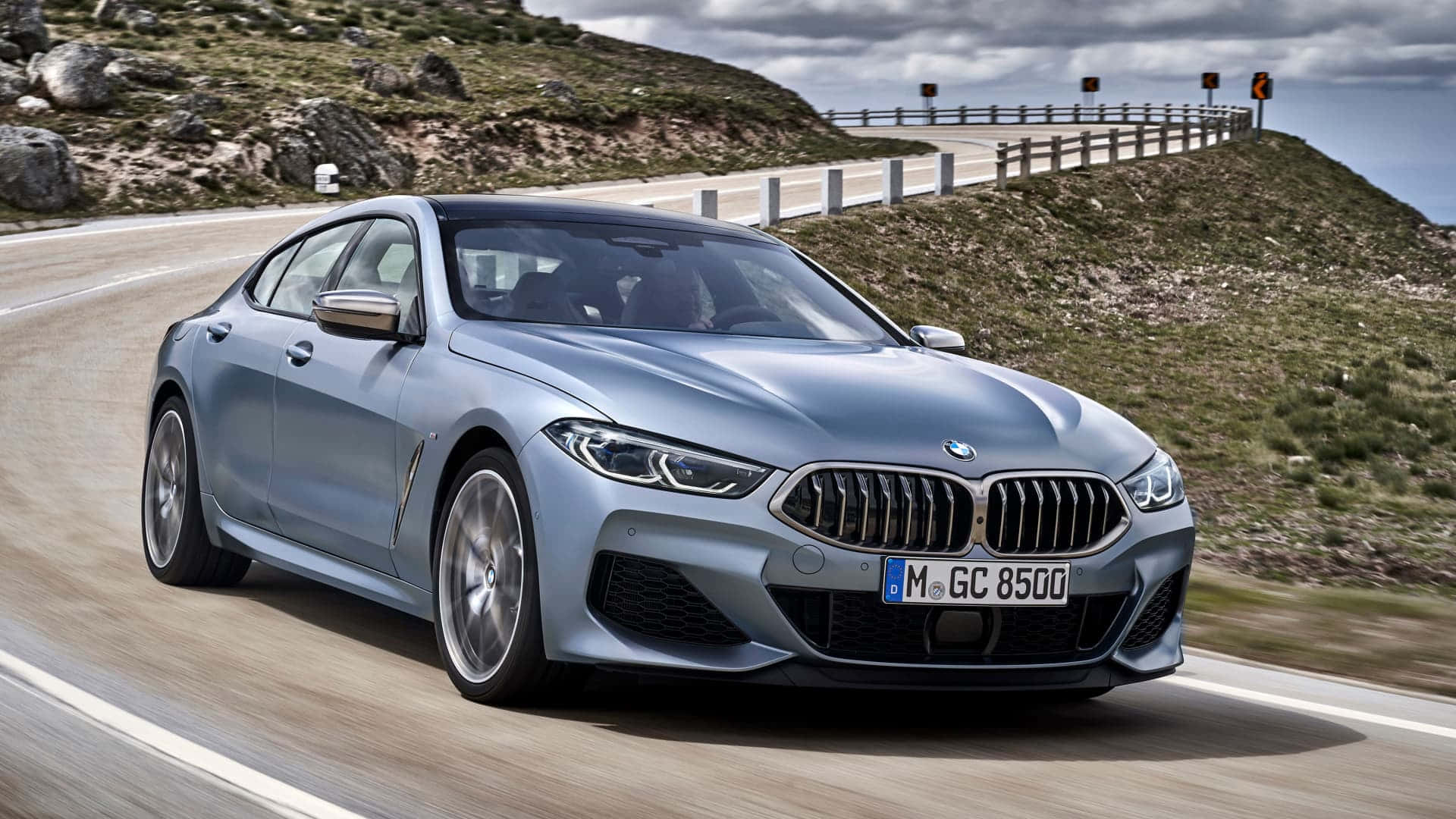 駕駛BMW，享受敞闊道路 壁紙 — 一輛時尚的銀色BMW 8系轎跑車在陰天下行駛在蜿蜒的山路上。