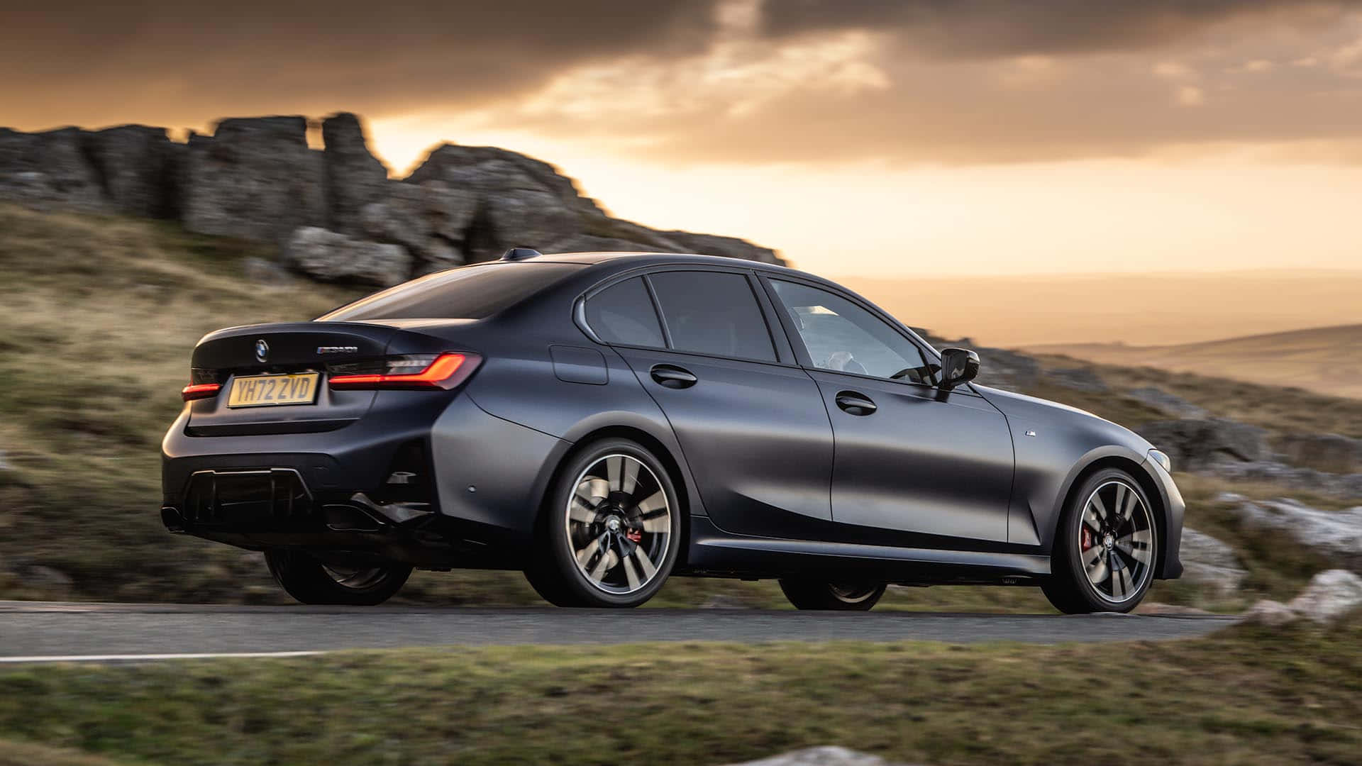 駕駛最新BMW，暢遊都市生活 壁紙 — 一輛時尚的啞光灰色BMW M3轎車在日落時分沿著蜿蜒的鄉村道路行駛，背景是岩石丘陵。