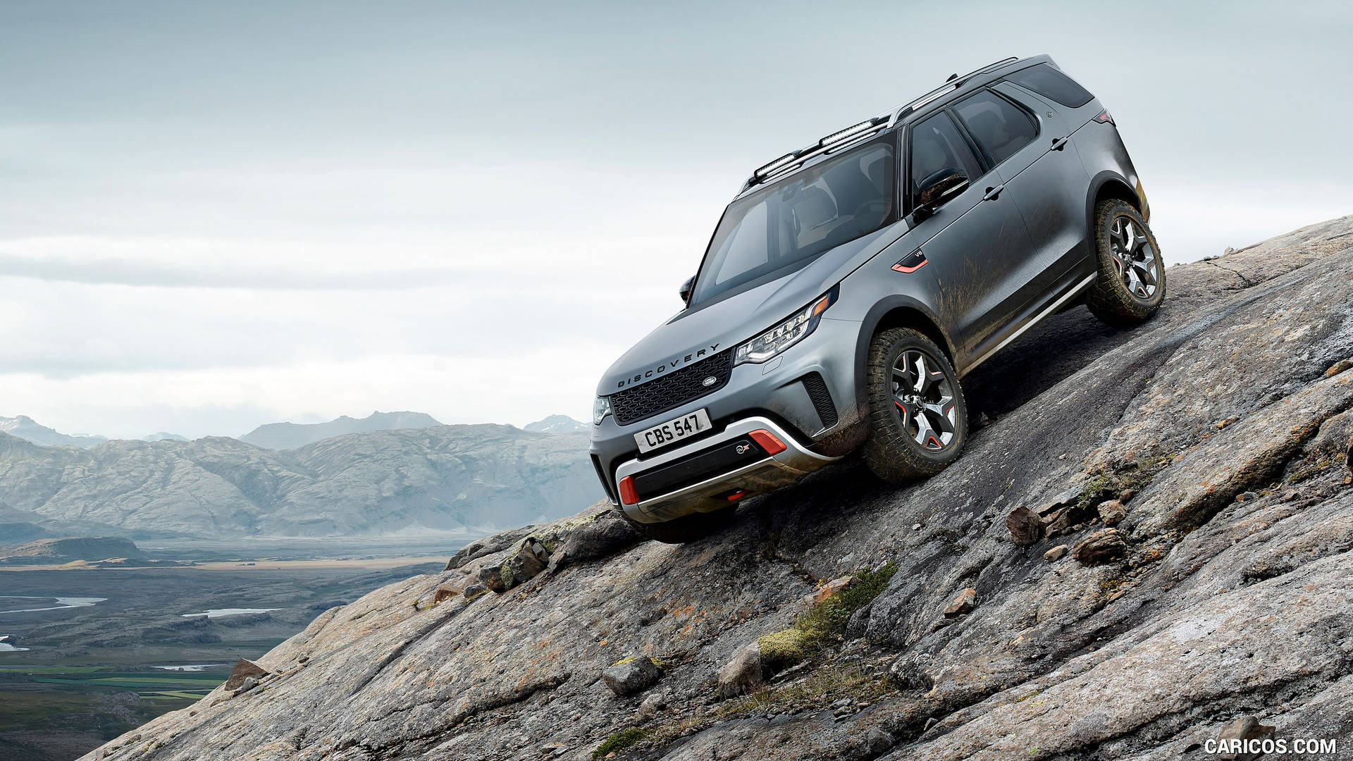 灰色 Land Rover Discovery 在陡峭岩石地形上攀爬，背景為山脈。