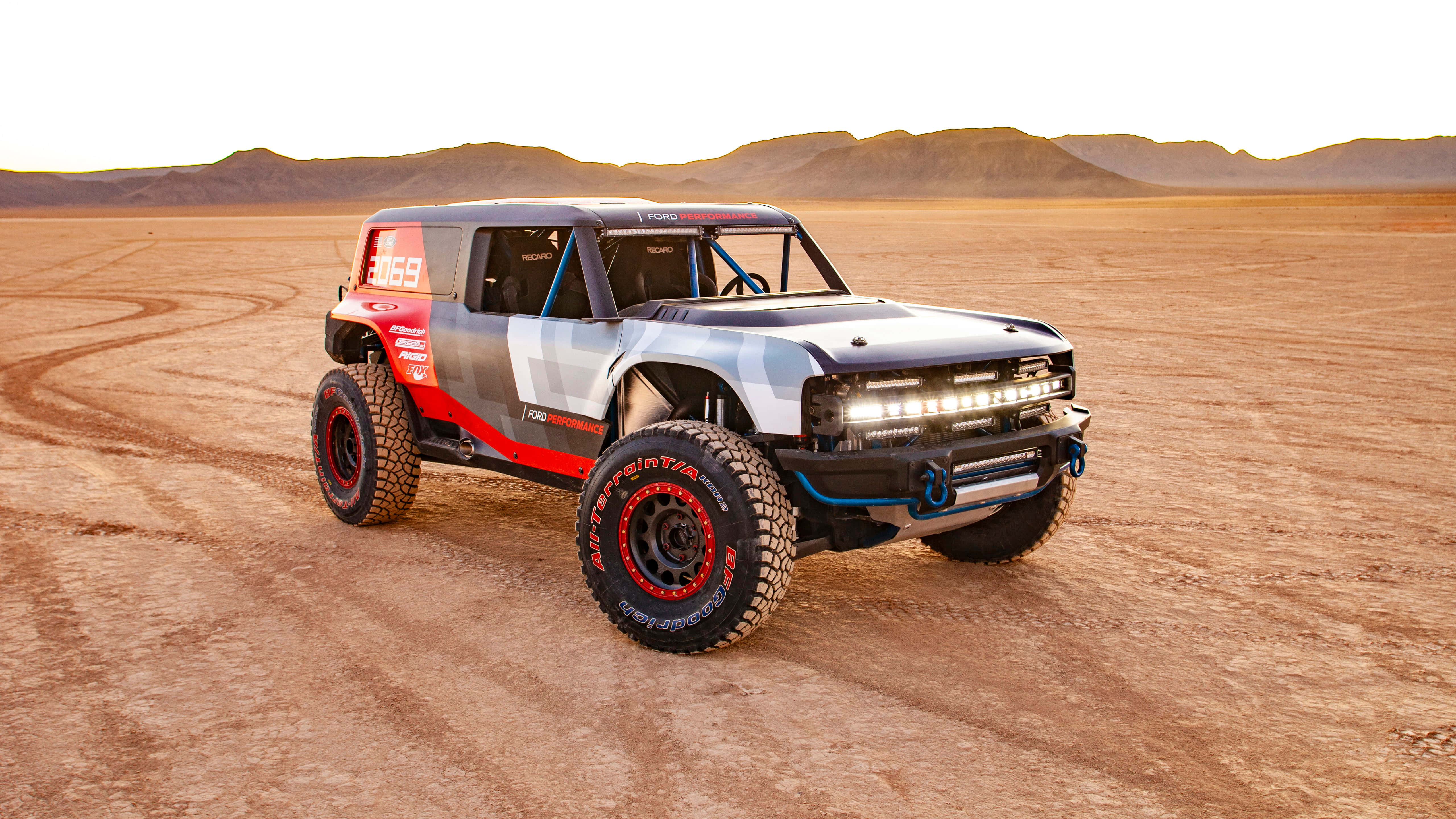 駕駛福特Bronco，征服越野冒險 壁紙 — 一輛堅固的福特Bronco賽車卡車，帶有紅色點綴和超大輪胎，在日落時分疾馳穿越廣闊的沙漠景觀。