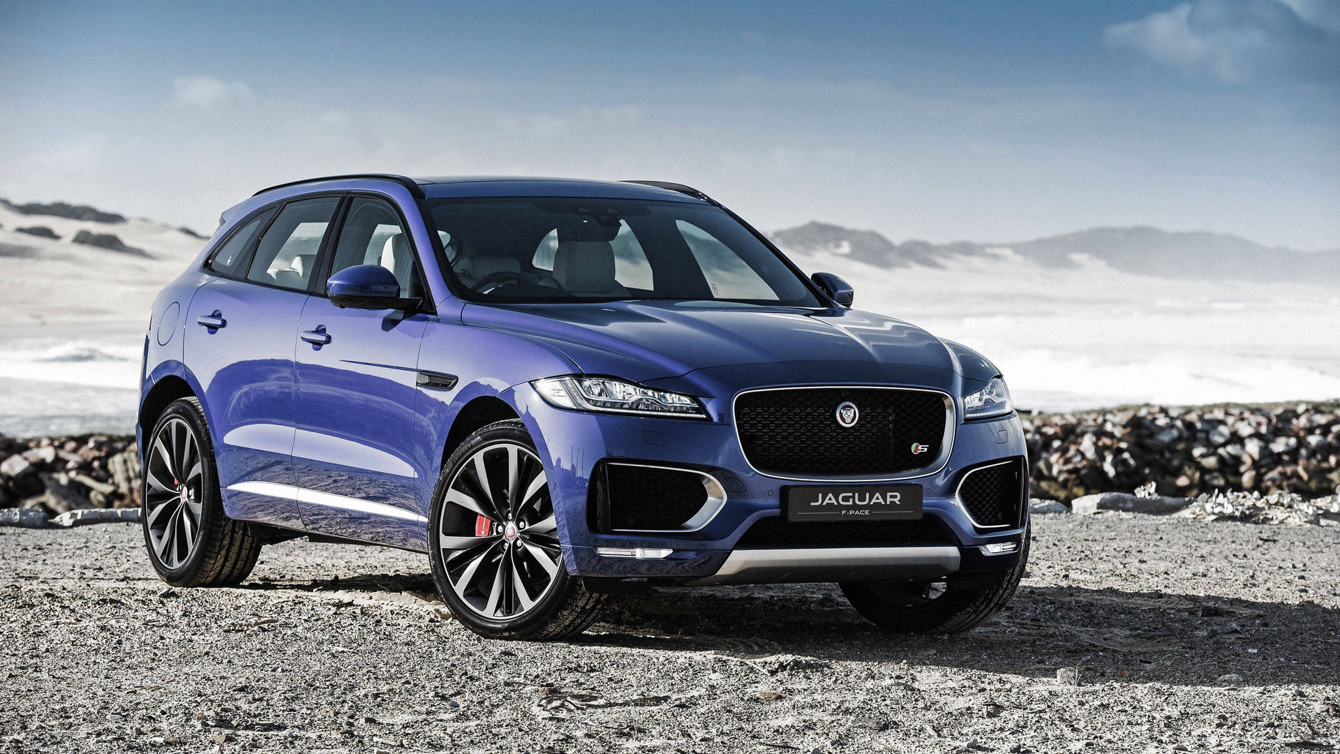 藍色Jaguar F-Pace SUV停泊在靠近雪山嘅岩石海灘上。