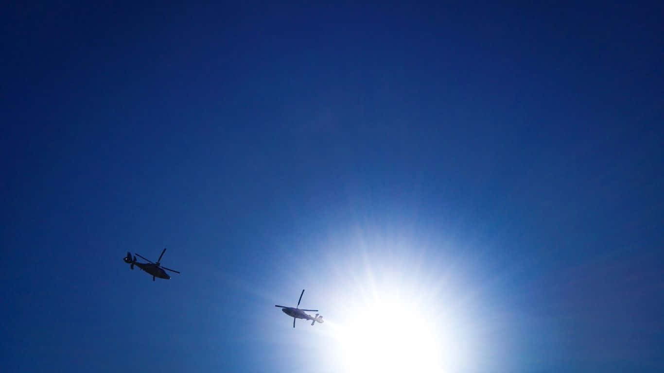 駕駛軍用直升機翱翔高空 壁紙 — 兩架直升機在明亮的太陽和深藍色天空的襯托下以剪影飛行。