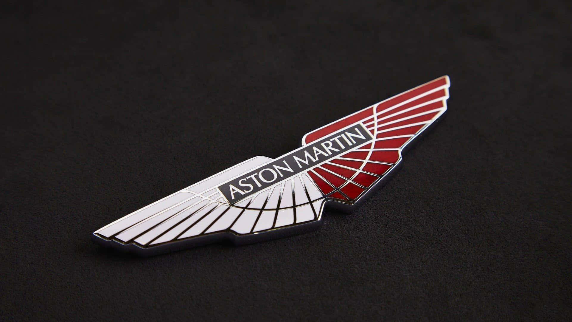 駕駛阿斯頓·馬丁，感受奢華的力量 壁紙 — 一個時尚的Aston Martin翼形標誌徽章，帶有紅色和銀色琺瑯，放置在磨砂黑色表面上。