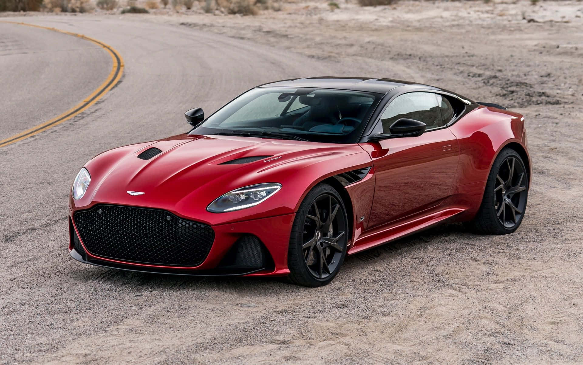 駕駛阿斯頓·馬丁，感受極致的尊榮與動力 壁紙 — 一架型格紅色Aston Martin DBS Superleggera停泊在蜿蜒的沙漠公路上，背景是崎嶇的地形。