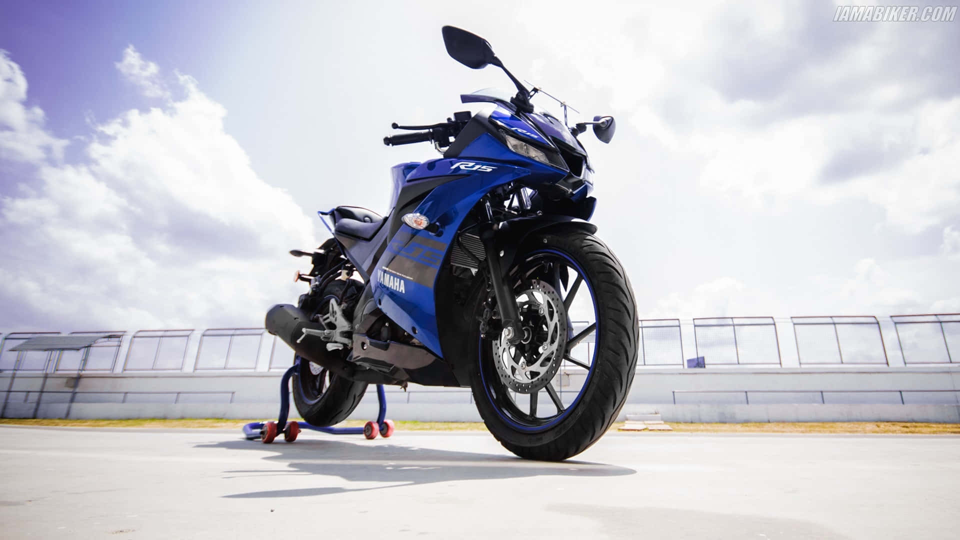 騎乘R15，盡享時尚風采 壁紙 — 一架鮮藍色的Yamaha R15電單車在晴朗天空和零星雲朵下立於支架上。