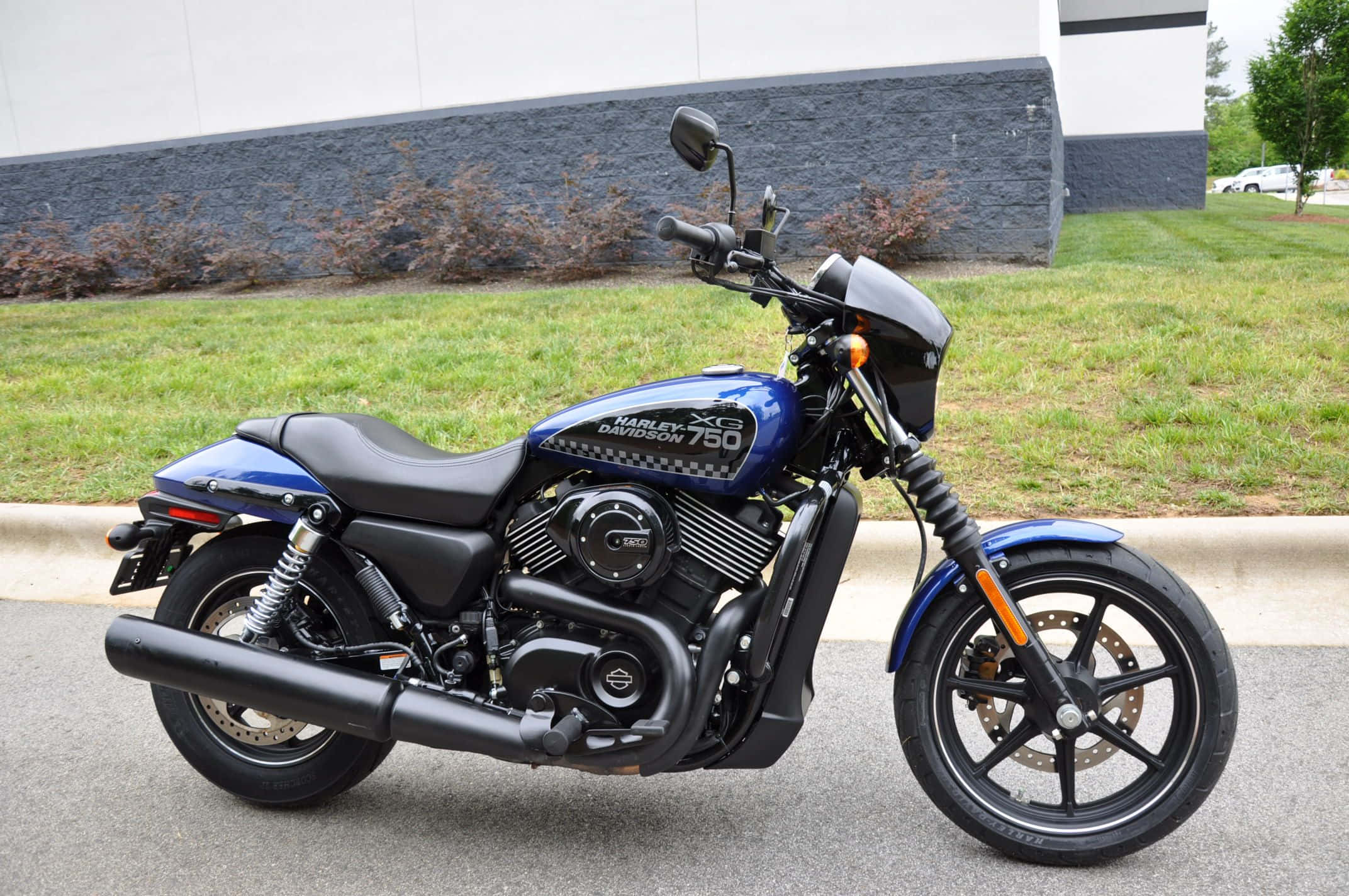 騎乘哈雷戴維森暢遊開闊道路 壁紙 — 一架時尚的藍色Harley-Davidson XG 750電單車停泊在行人路上，旁邊有草地和現代建築。