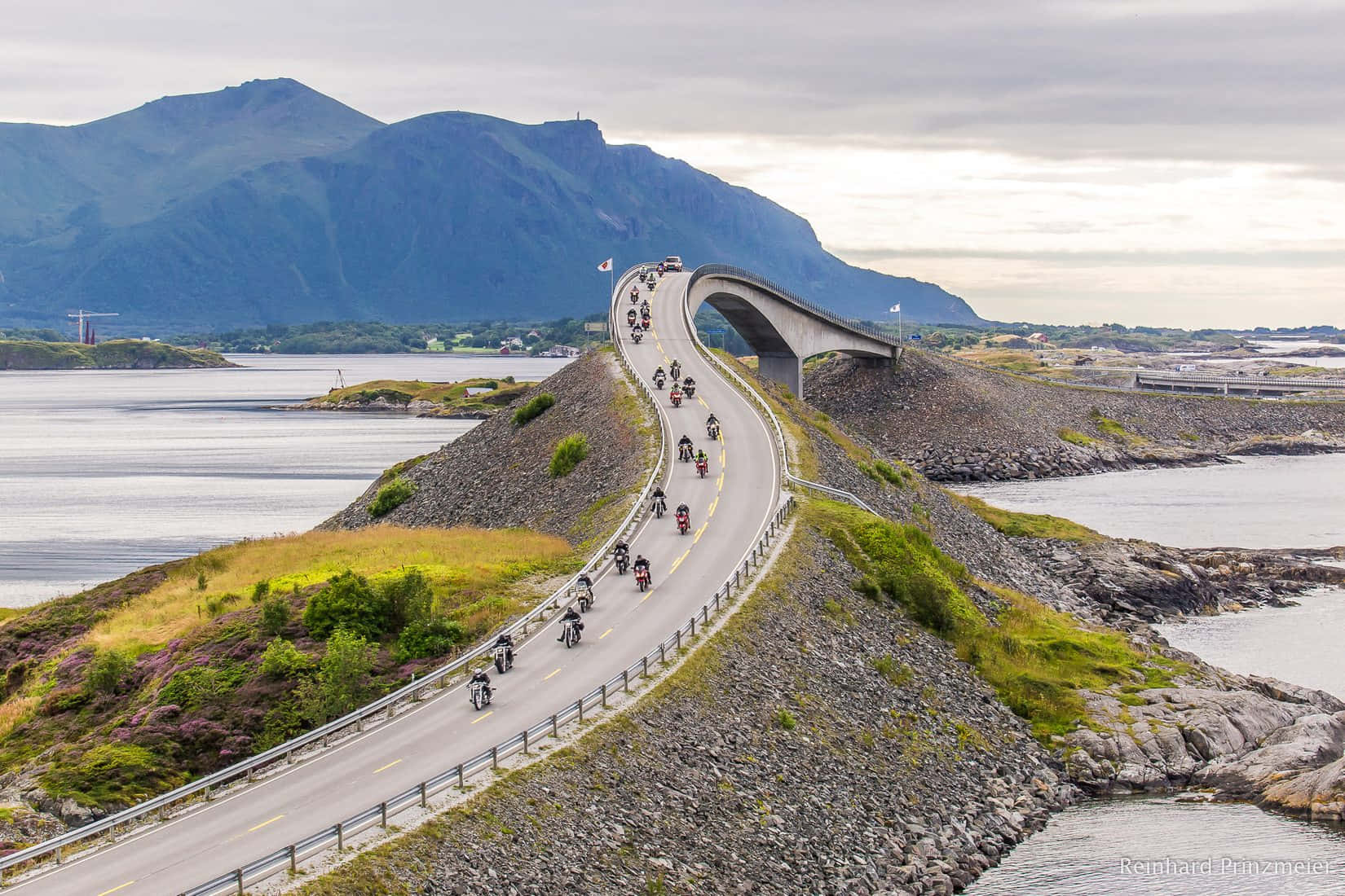 電單車手沿著風景優美的海岸道路行駛，背景有山脈和水域。