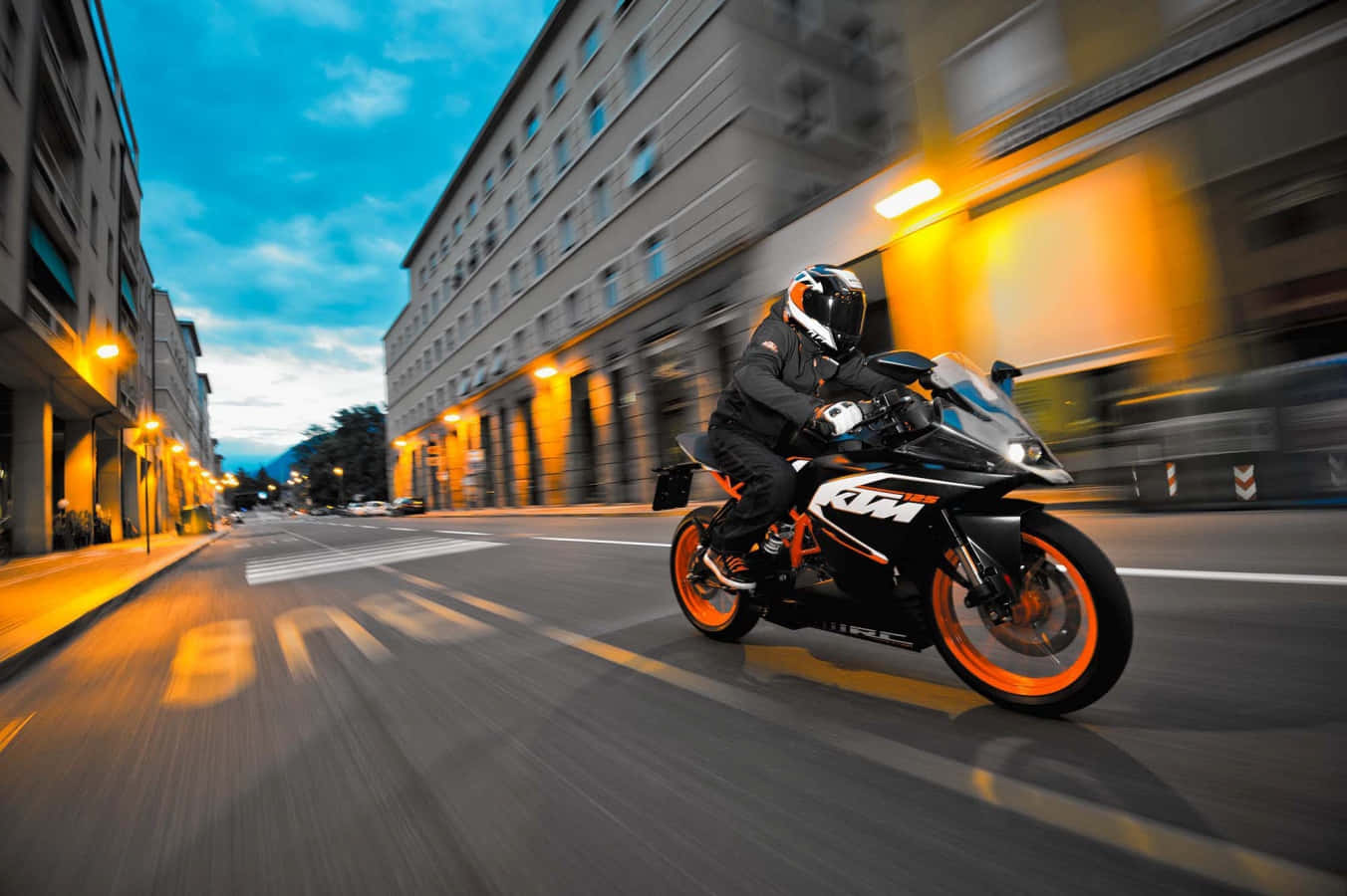 騎士駕駛KTM RC 125穿街過巷動態照片 壁紙 — 一名騎士騎著黑橙色的KTM運動電單車，在黃昏時分伴隨著模糊的動態和溫暖的路燈，飛馳在城市的街道上。