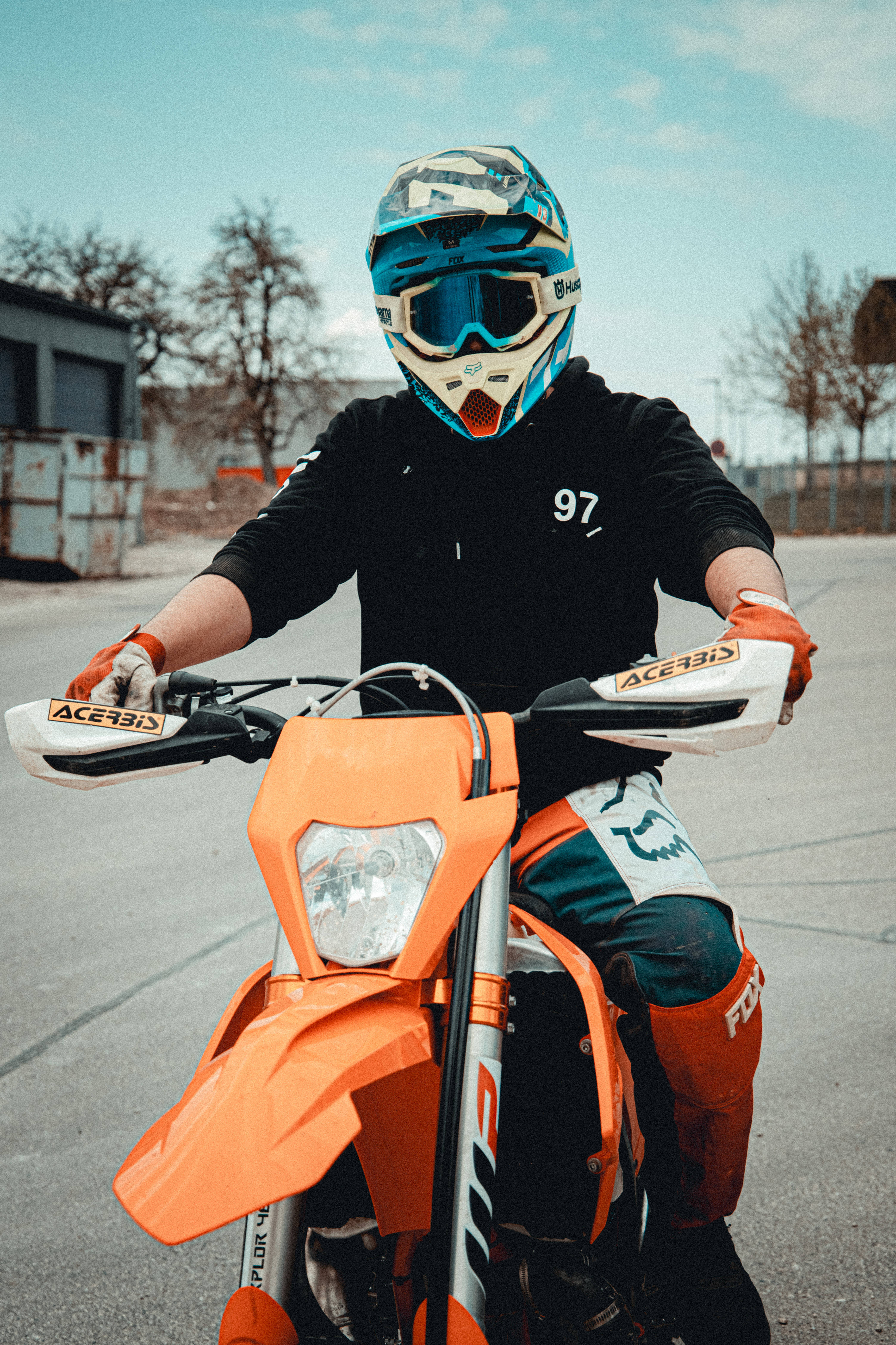 騎士駕駛橙色KTM機車 壁紙 — 一位身穿全套裝備的越野電單車手，在晴朗天空下的瀝青地上，握著一輛亮橙色越野車的把手。