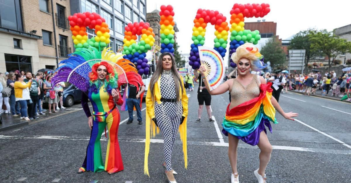 三位變裝表演者穿著彩虹服裝，手持拼出PRIDE的氣球字母遊行。
