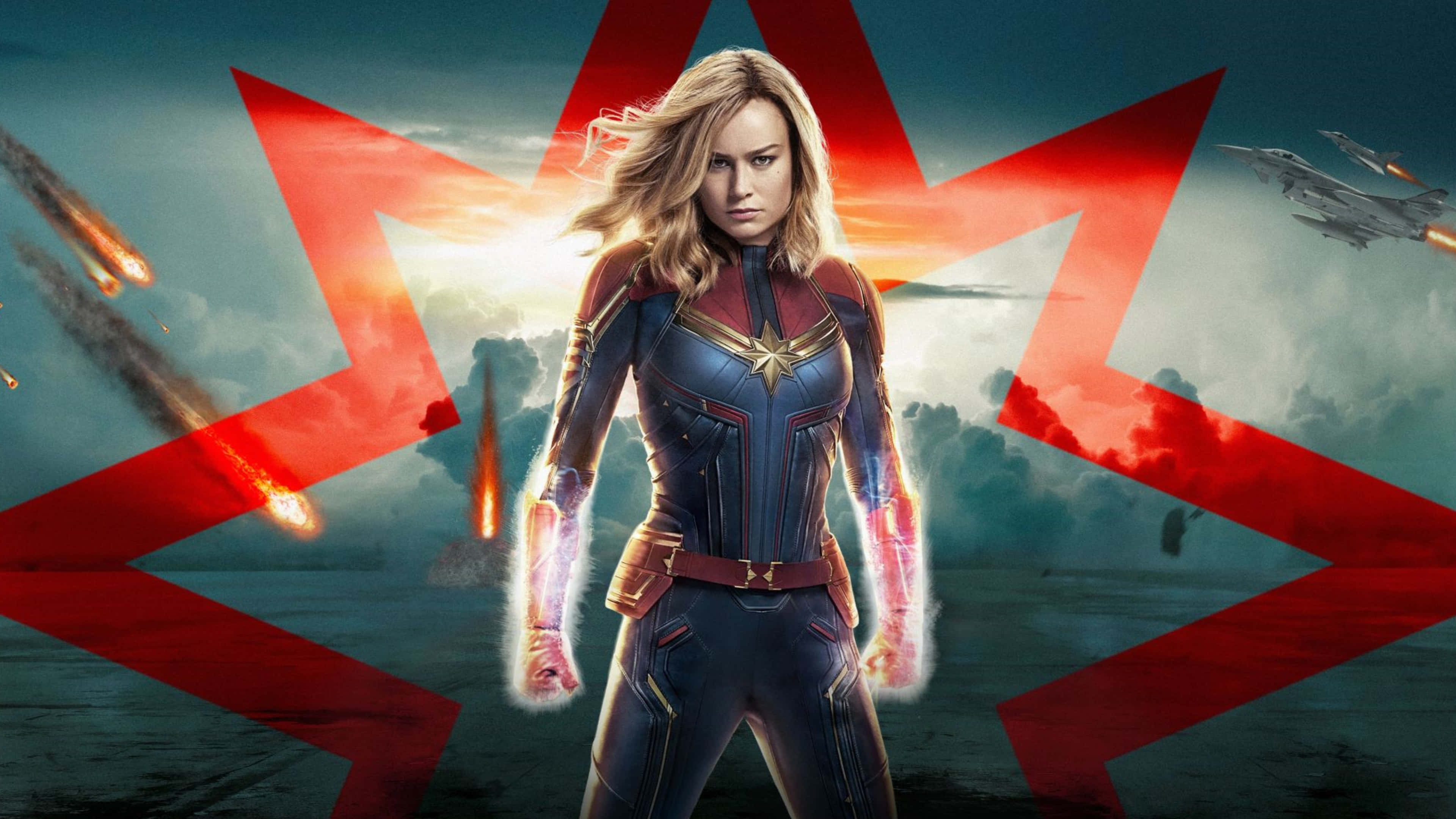 4K Marvel 壁紙 桌布 壁紙 — 驚奇隊長挑釁地站著，拳頭發光，流星劃過，戰鬥機在戲劇性的日落天空下飛行，天空由大膽的紅星框住。