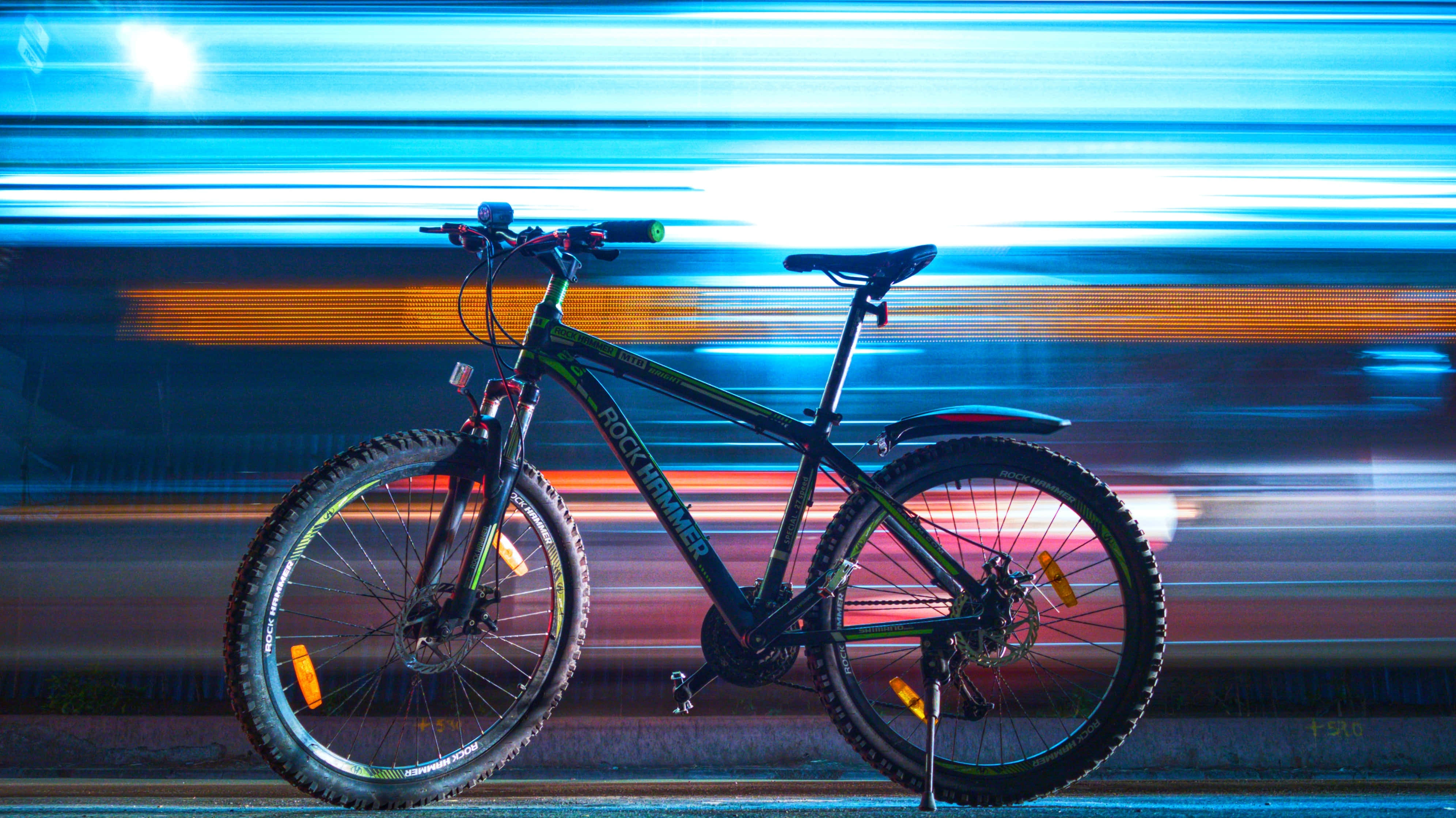 驚艷Rock Hammer硬尾山地車背景 壁紙 — 一輛山地單車在夜晚的霓虹城市景觀前靜止，帶有藍色和橙色的光跡。