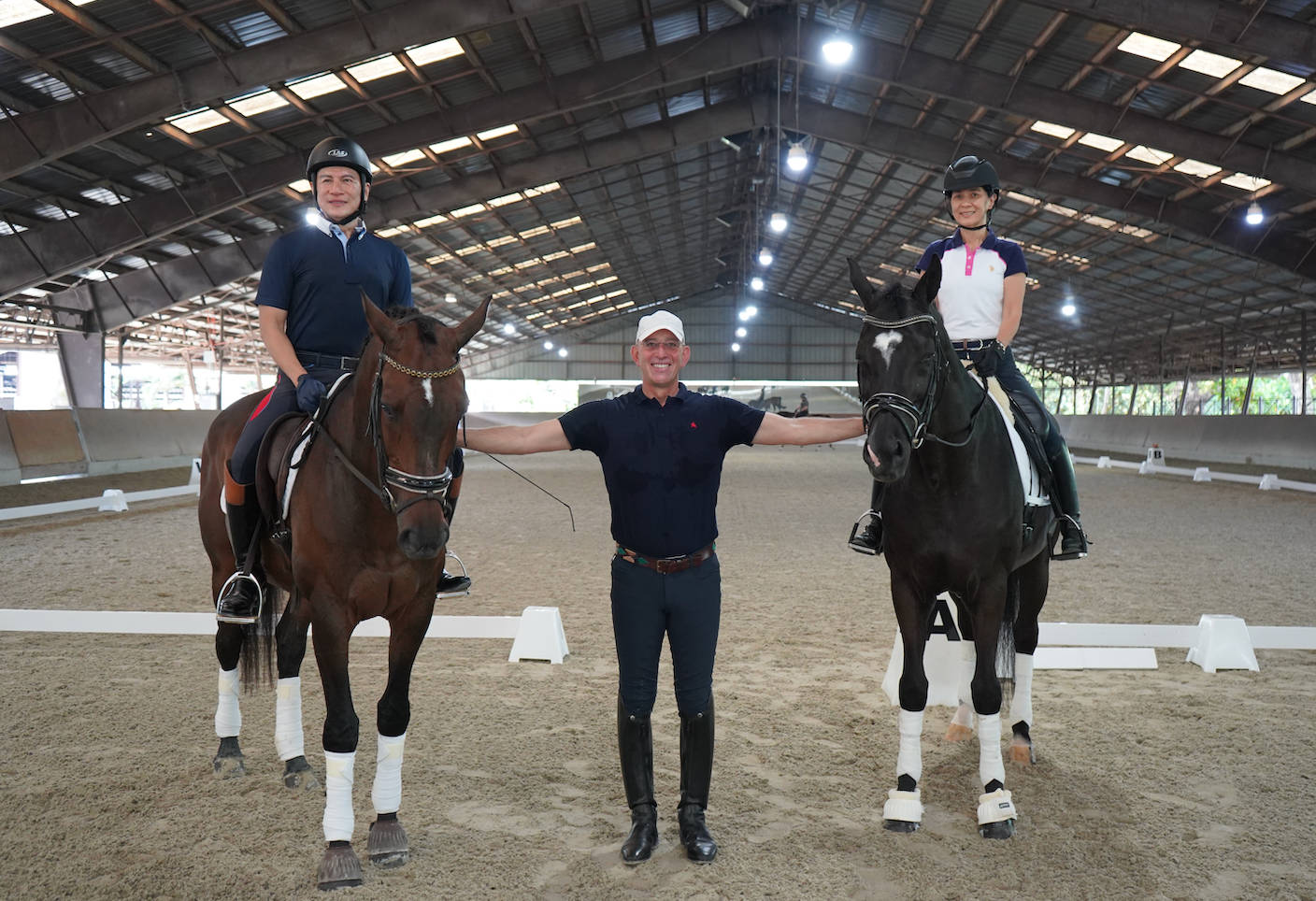 驚艷馬術教練同時指導兩位騎手 壁紙 — 兩位騎手騎著馬，在一個有高天花燈的室內場地中，站在一位微笑的男士兩旁。