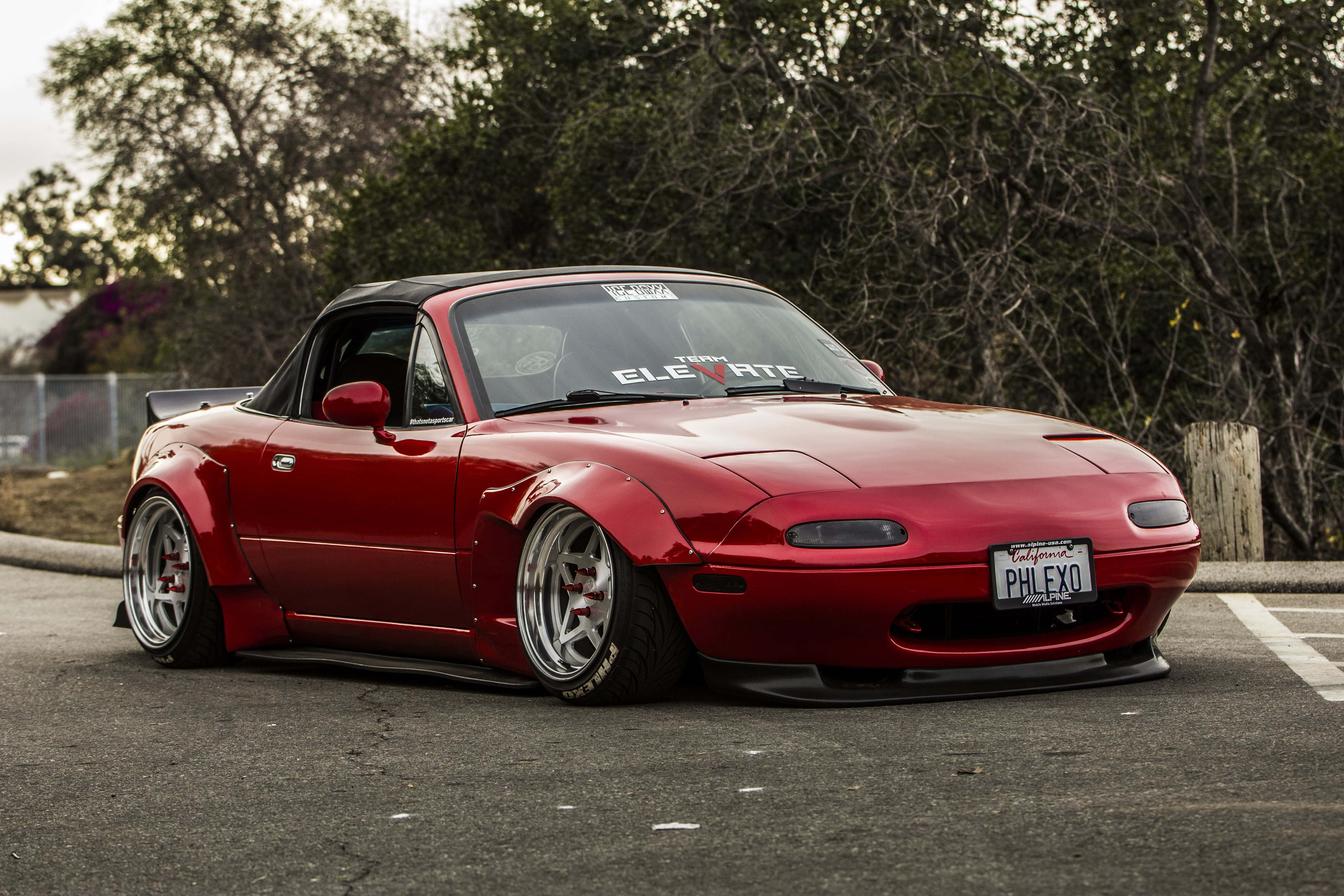 驚豔的紅色馬自達MX-5 Miata 壁紙 — 一輛光澤紅色Mazda Miata開篷跑車，配備寬體套件和訂製輪圈，停泊在樹木環繞的停車場。