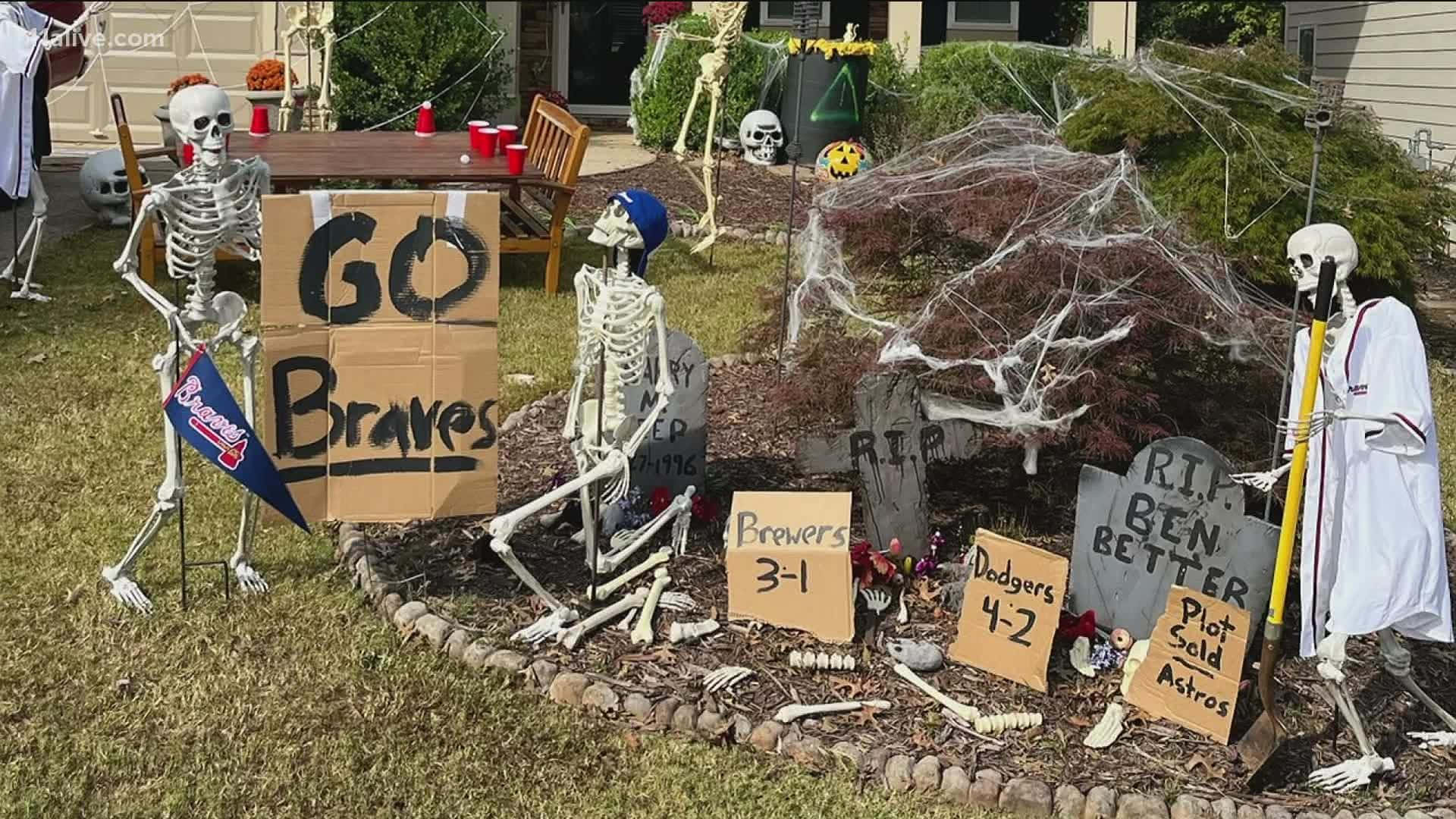 Halloween Yard Decorations 桌布 壁紙 — 骷髏人物穿著萬聖節服裝，在節日庭院佈置中愉快地展示棒球隊標誌和墓碑。