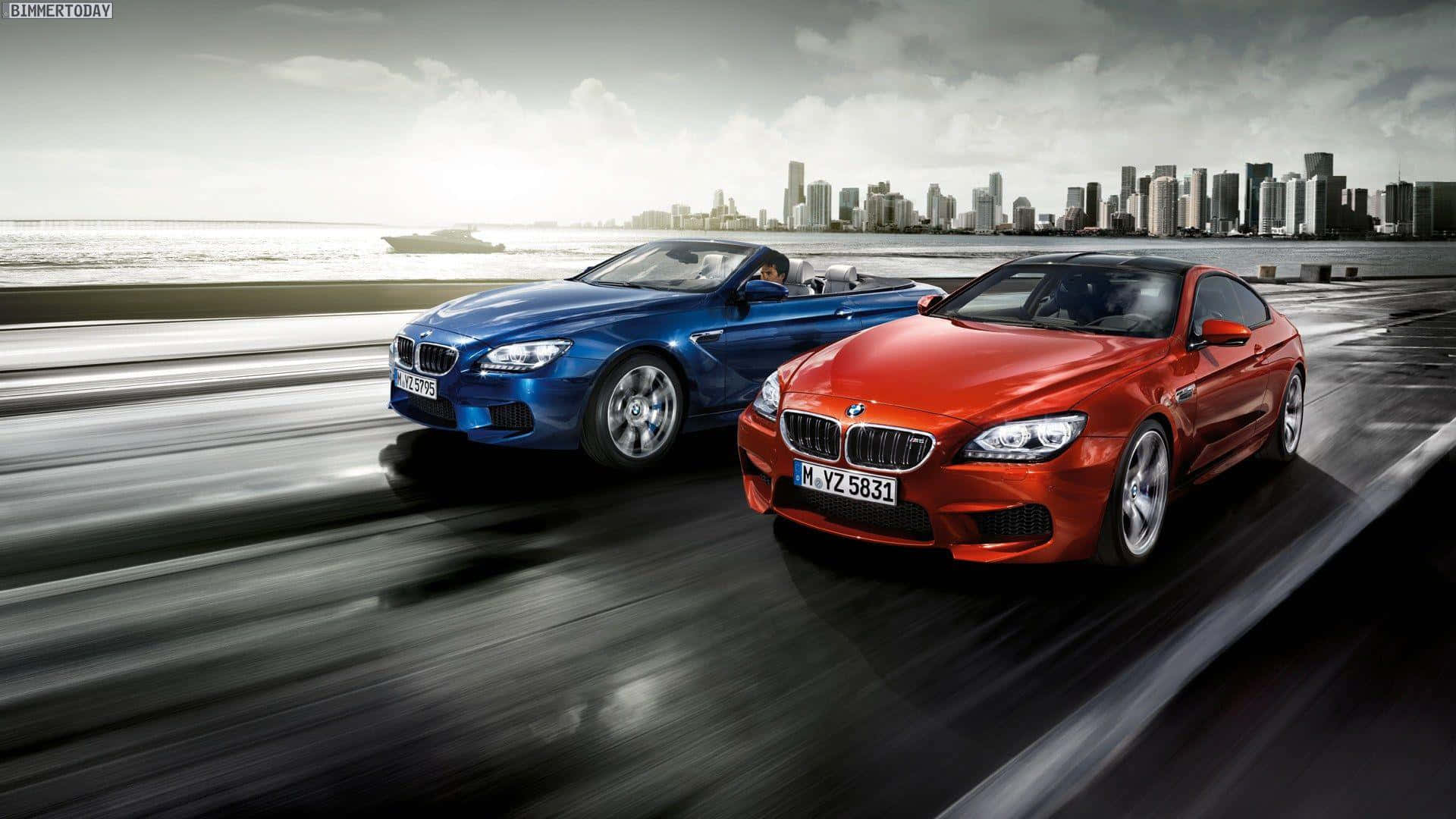 體驗 1920x1080 M系列的震撼視覺 壁紙 — 兩輛流線型BMW跑車沿著濕漉漉的海岸高速公路疾馳，背景是城市天際線和海洋。