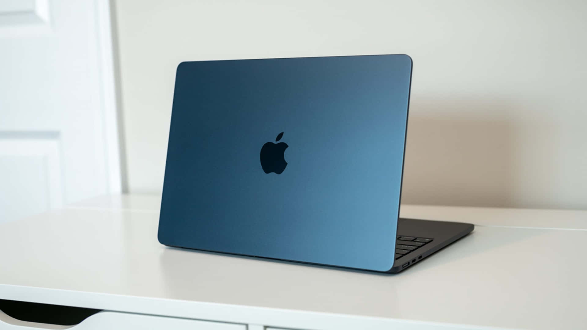 體驗 Apple Macbook 的卓越清晰度與鮮豔色彩 壁紙 — 一部時尚的藍色 MacBook 打開放在白色桌子上，柔和自然光突顯其啞光質感。