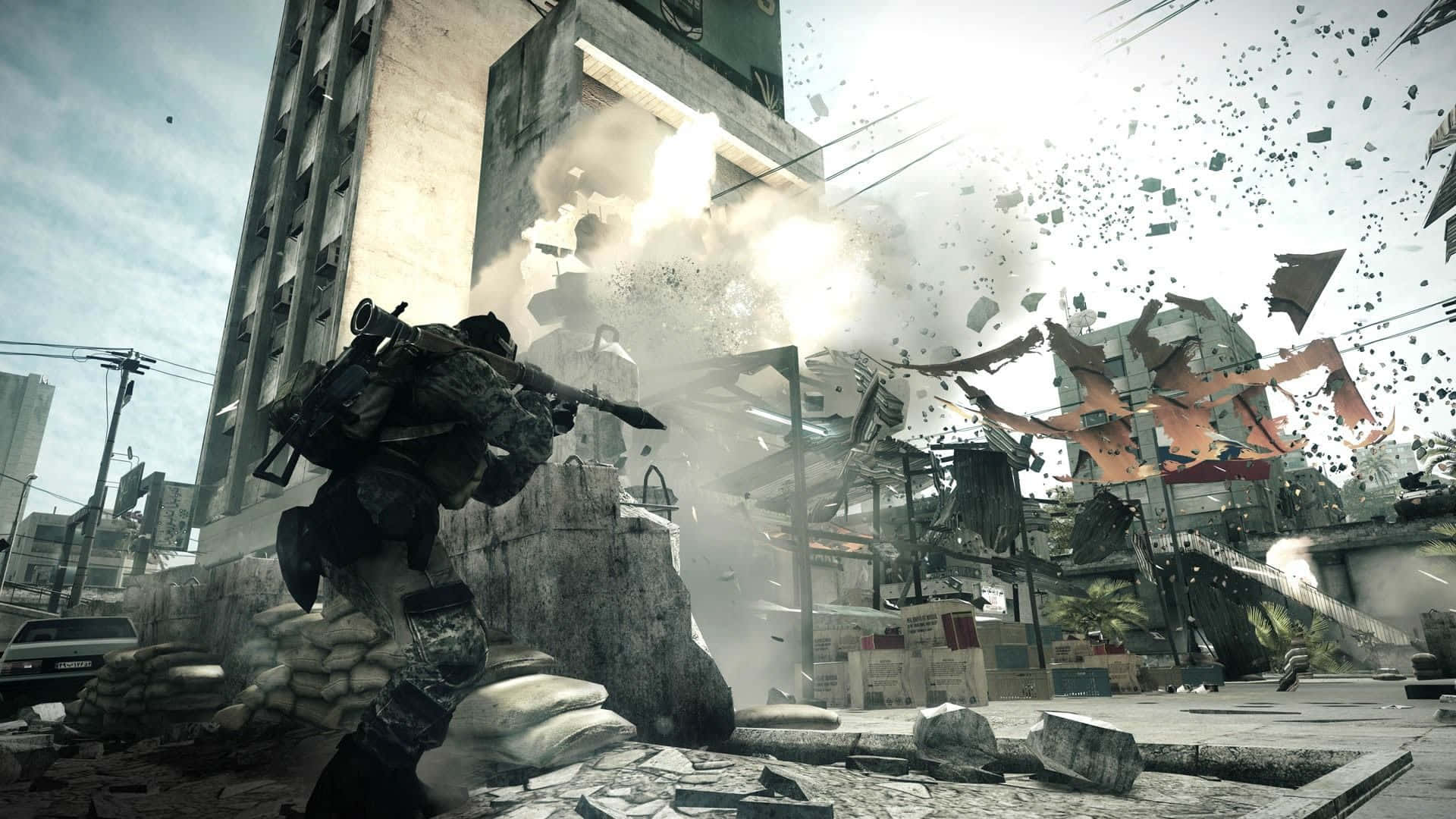 體驗《Battlefield 3》的激烈戰鬥 壁紙 — 一名士兵在沙包後方掩護，爆炸撕裂了廢墟般的城市街道，碎片四處飛散。