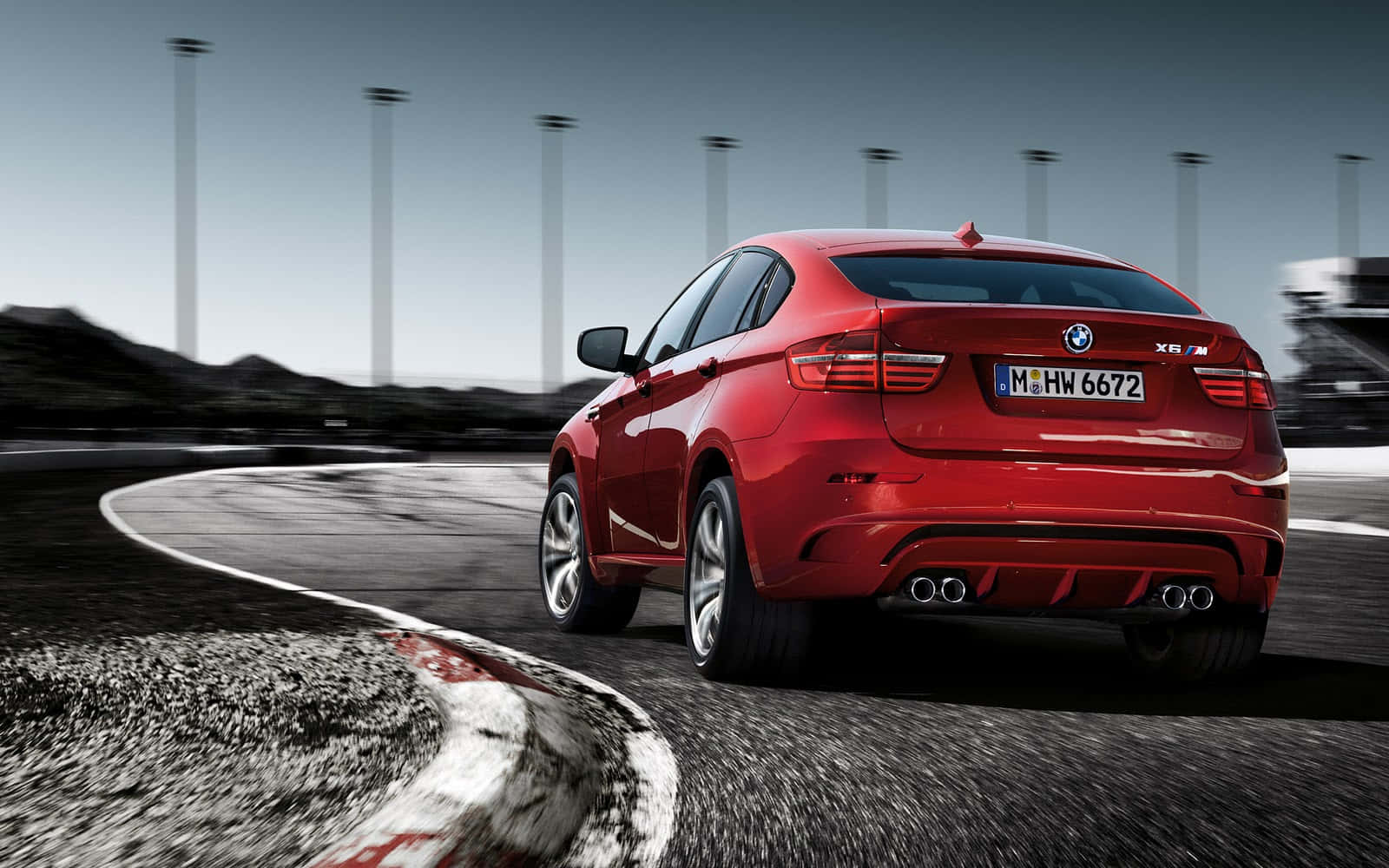 體驗 BMW X6 M 的原始動力 壁紙 — 一輛鮮紅色的BMW X6 M在賽道彎位高速飛馳，瀝青路面呈現動態模糊效果，背景有高聳的燈柱。