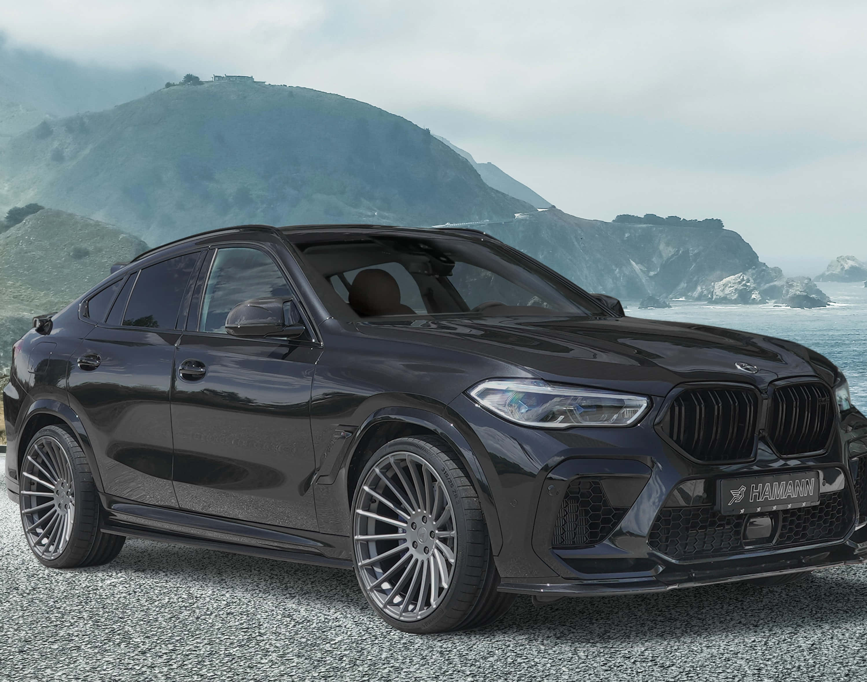 體驗 BMW X6 M 的極致動力 壁紙 — 一架型格黑色Hamann改裝BMW X6停泊在海邊碎石上，背景有霧氣籠罩的綠色山丘。