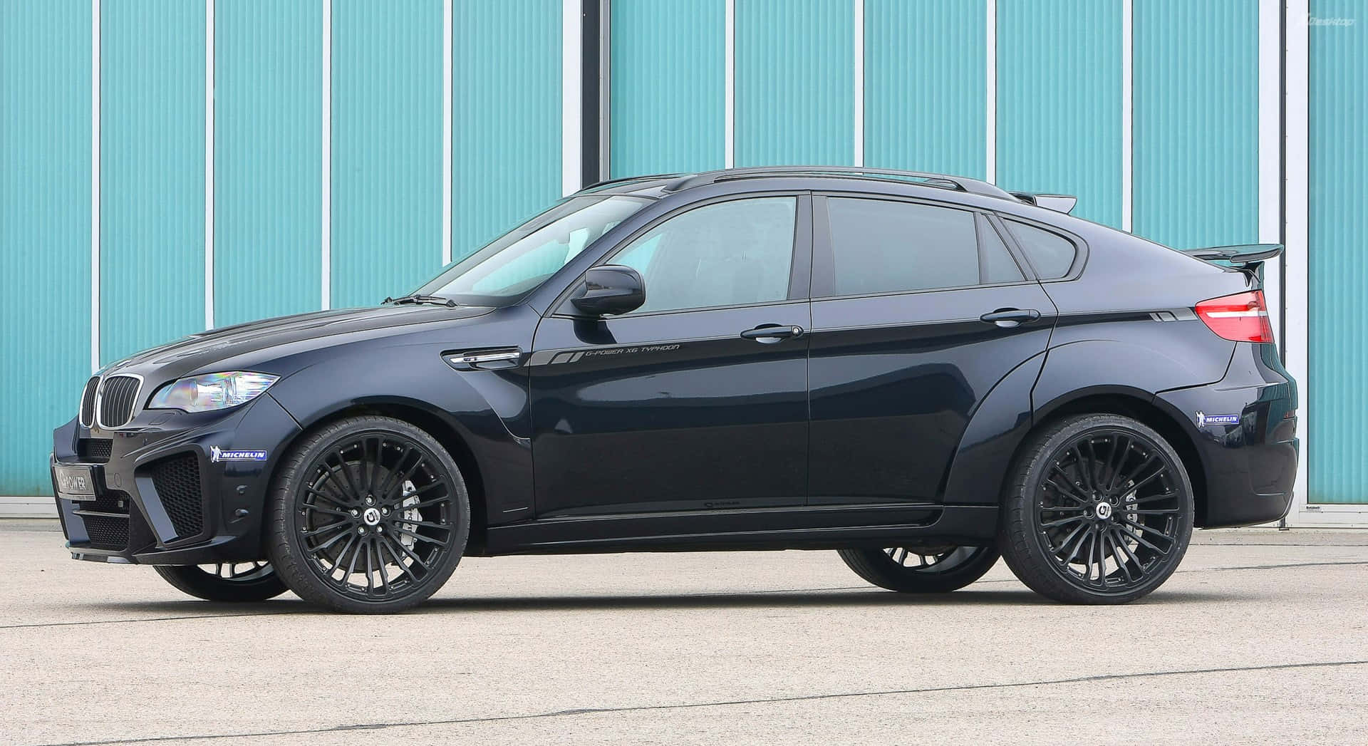 體驗 BMW X6 M 的極致駕馭快感 壁紙 — 一架時尚黑色BMW X6M跑車停泊在青綠色波紋金屬牆前。