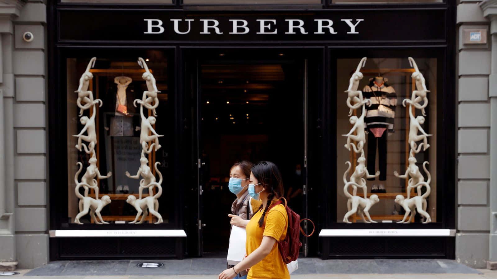 體驗 Burberry 的經典英國時尚 壁紙 — 兩名戴著口罩的行人經過一間Burberry商店，其櫥窗內有引人注目的白色猴子雕塑。