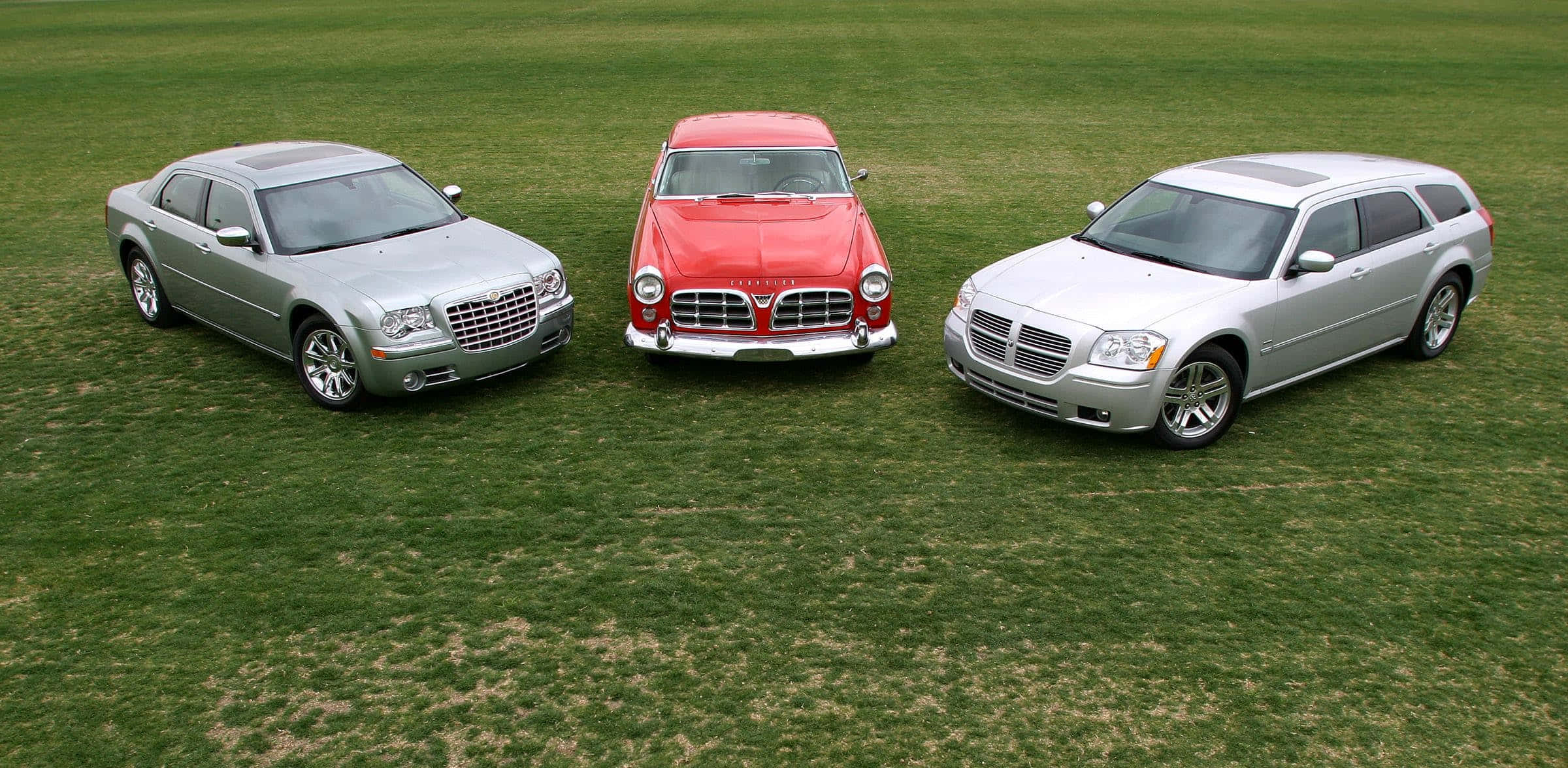 體驗 Chrysler 帶來的奢華與舒適 壁紙 — 三輛不同的克萊斯勒汽車，銀色、紅色和銀色，在明亮的日光下並排停放在茂密的綠色草坪上。