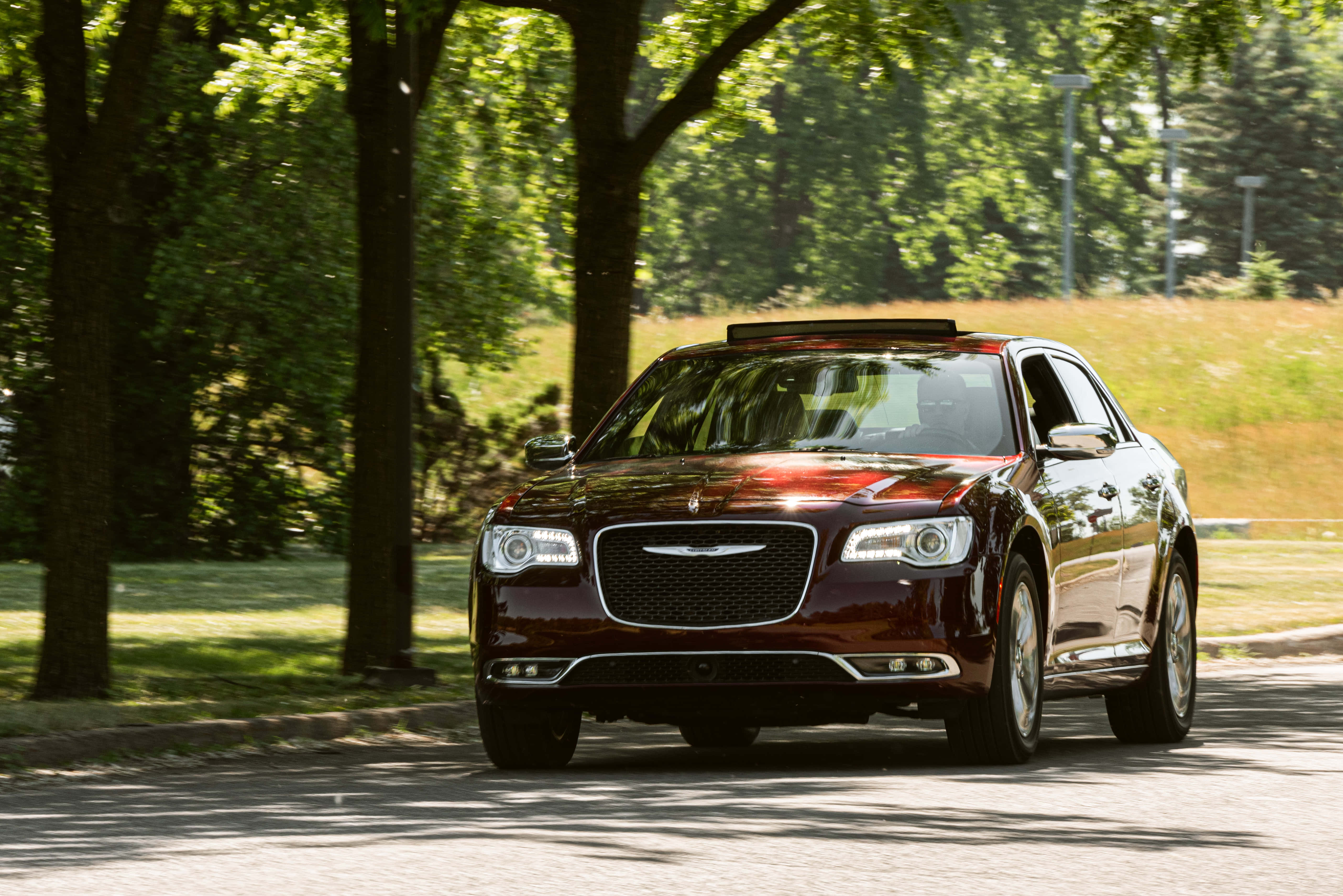 體驗 Chrysler 系列的奢華魅力 壁紙 — 一輛光亮的栗色豪華轎車在明亮的夏日陽光下行駛在一條綠樹成蔭的道路上，投下斑駁的陰影。