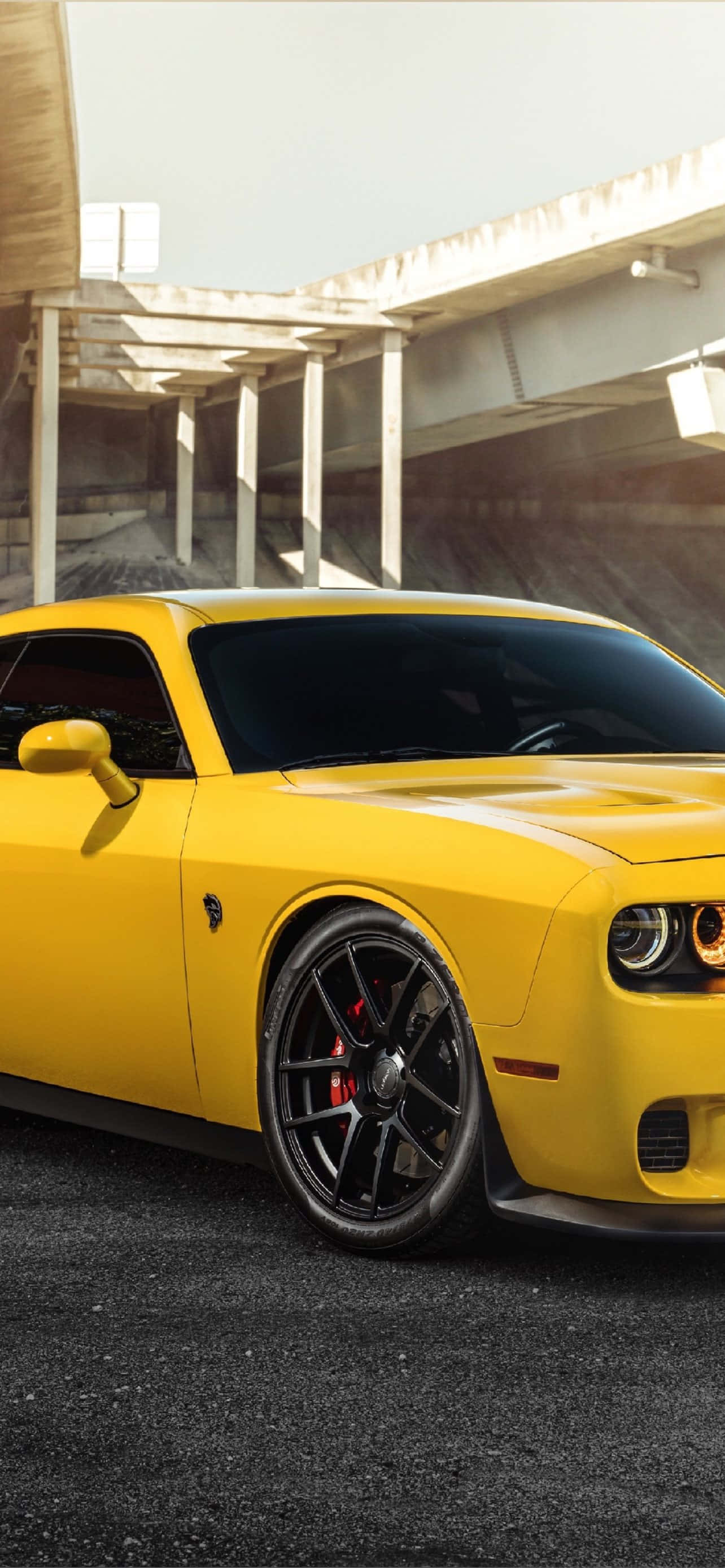體驗 Hellcat iPhone 的無與倫比速度與效能 壁紙 — 一輛鮮黃色的道奇挑戰者跑車停泊在混凝土天橋下，配備黑色輪圈和紅色制動卡鉗。