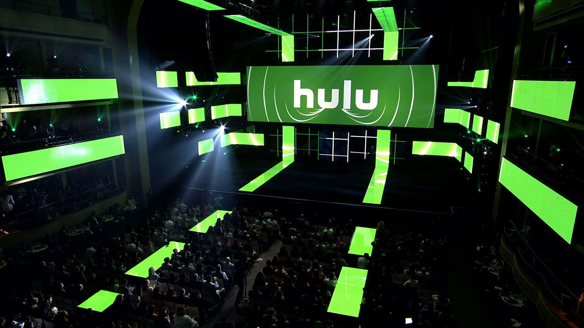 體驗 Hulu 及其無限可能 壁紙 — 一個大型禮堂，坐滿了觀看舞台的人，舞台沐浴在展示Hulu標誌的鮮豔綠色燈光中。