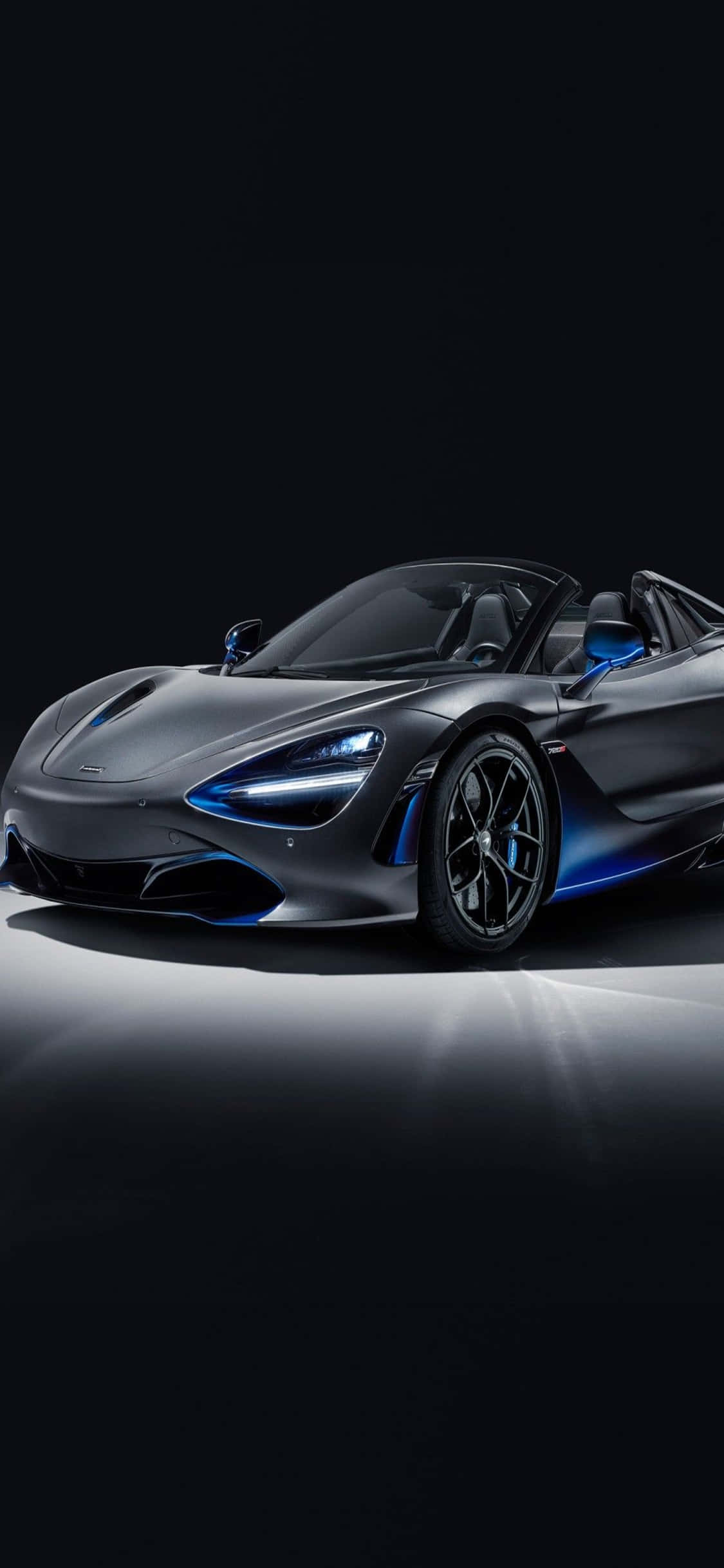 體驗 iPhone Xs 與 McLaren 720S 無與倫比的強大動力 壁紙 — 一輛時尚的啞光灰色McLaren 720S Spider，帶有發光的藍色飾邊和車頭燈，在黑色工作室背景下亮起。