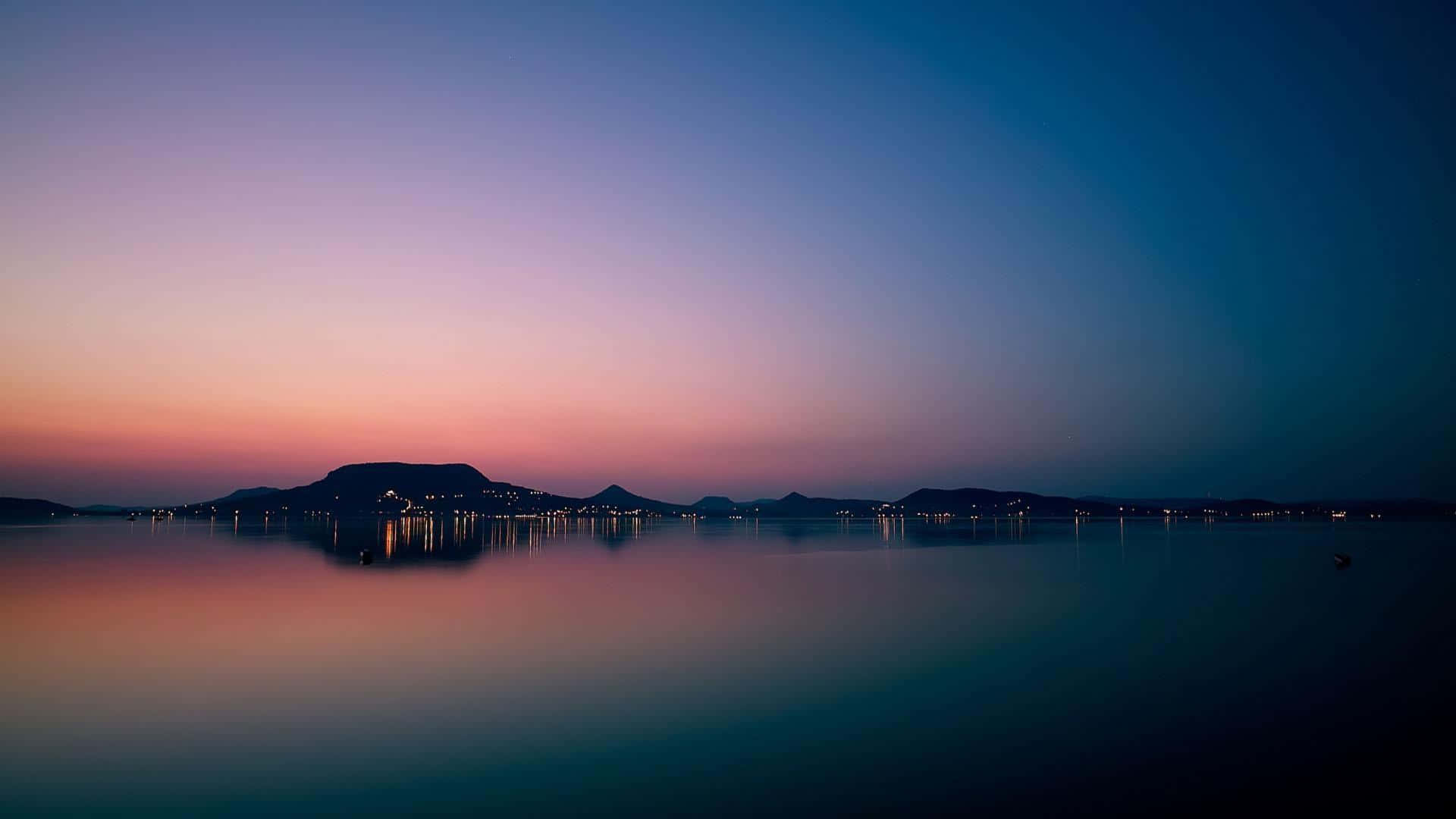 黃昏時平靜的湖泊反射出紫色和粉紅色的天空，遠方有山丘和城市燈光。