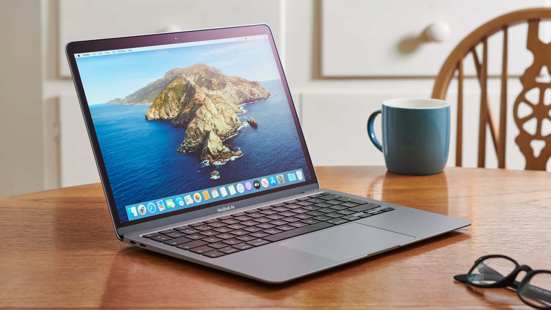 體驗 MacBook 的強大效能與便攜性 壁紙 — 一部時尚的銀色 MacBook Air 打開放在木桌上，顯示充滿活力的海岸島嶼壁紙，旁邊有一個藍色杯子和眼鏡。