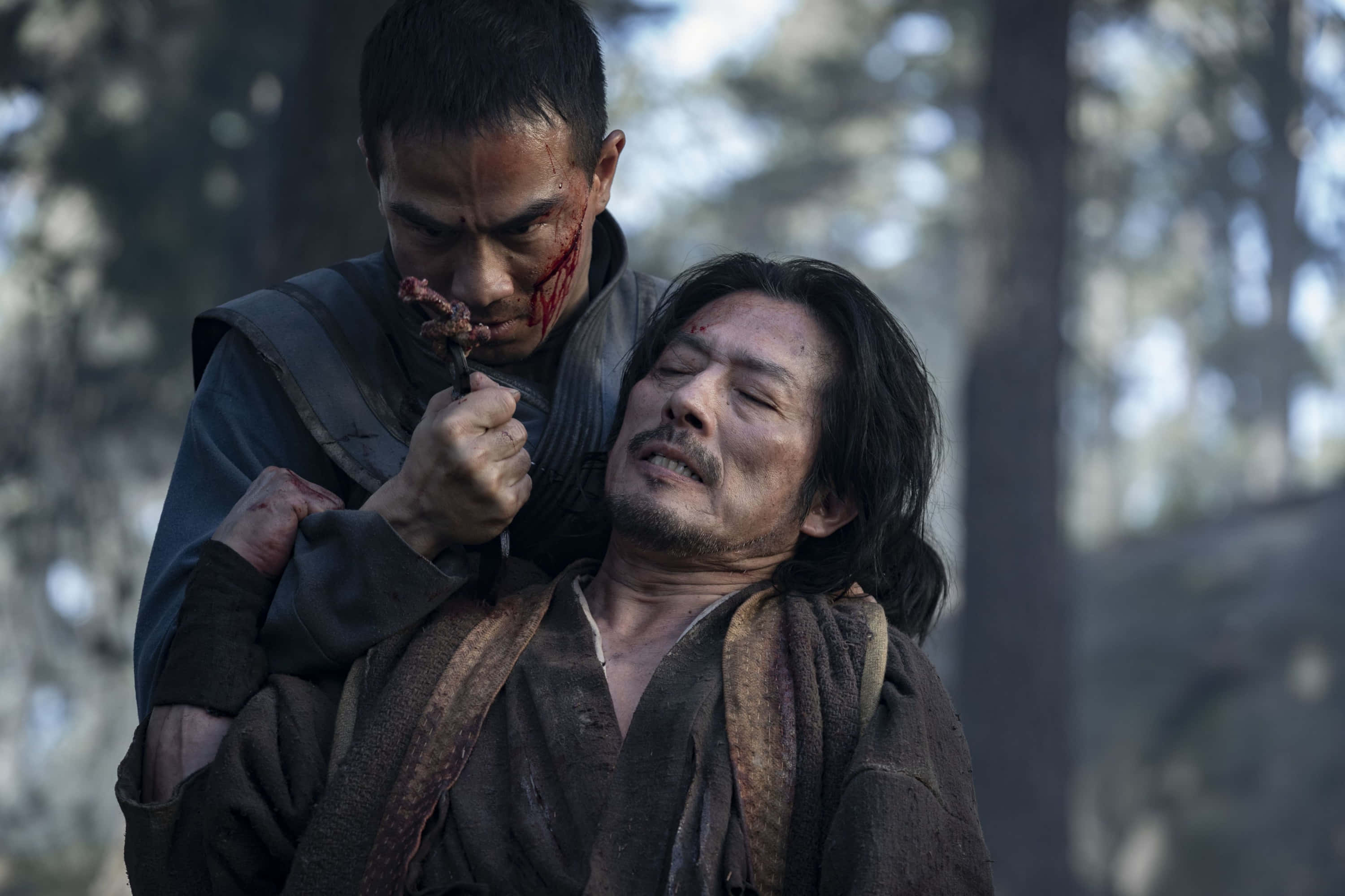 體驗《真人快打 2021》的刺激與戲劇性 壁紙 — 一個血跡斑斑的戰士在森林中，緊張對峙中將刀架在另一個男人的喉嚨上。
