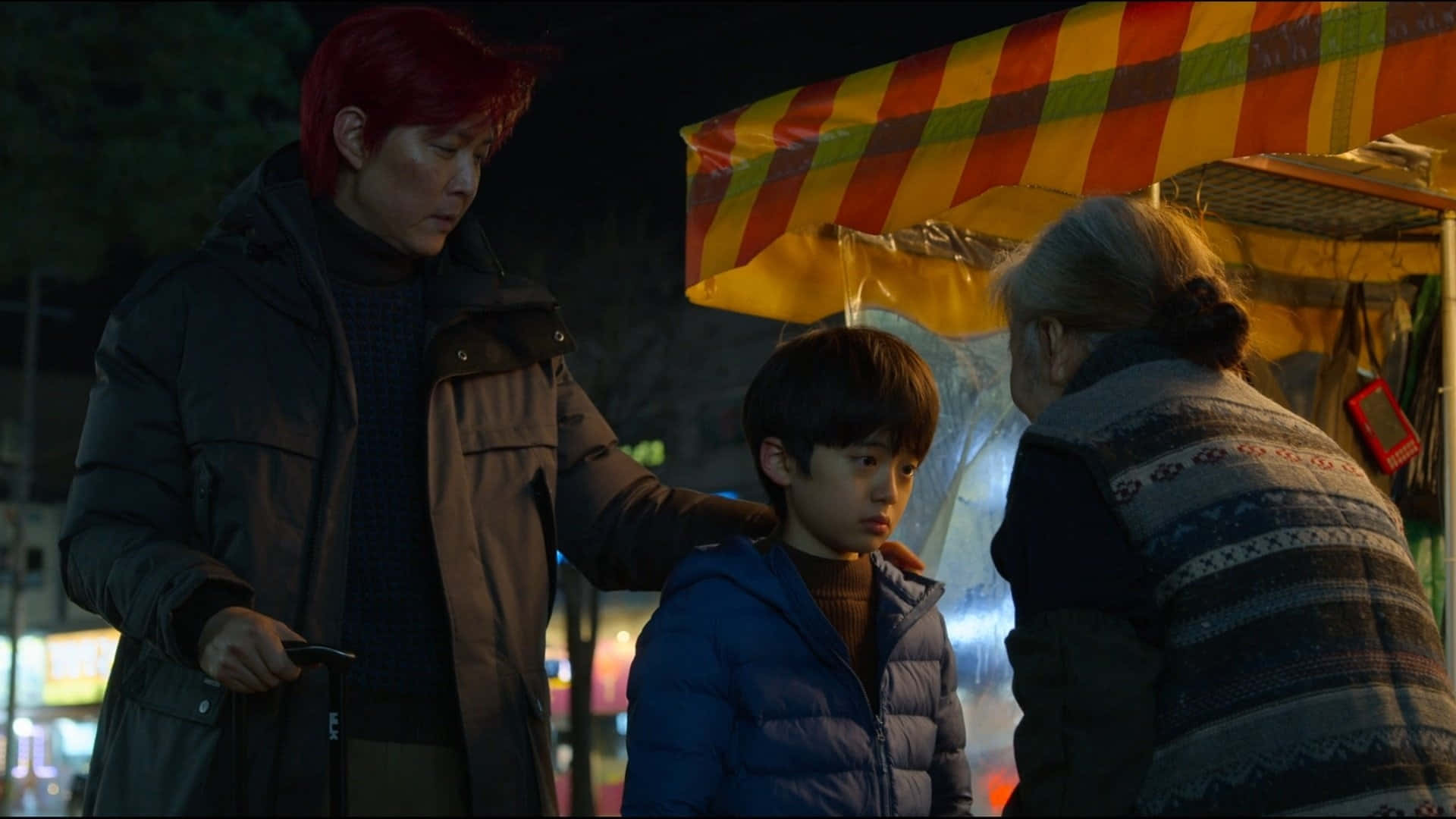 體驗《魷魚遊戲》Zoom的刺激 壁紙 — 一個紅髮男人和一個穿藍色外套的男孩站在一位年長女士附近，在夜燈下的一個色彩繽紛條紋美食攤檔。