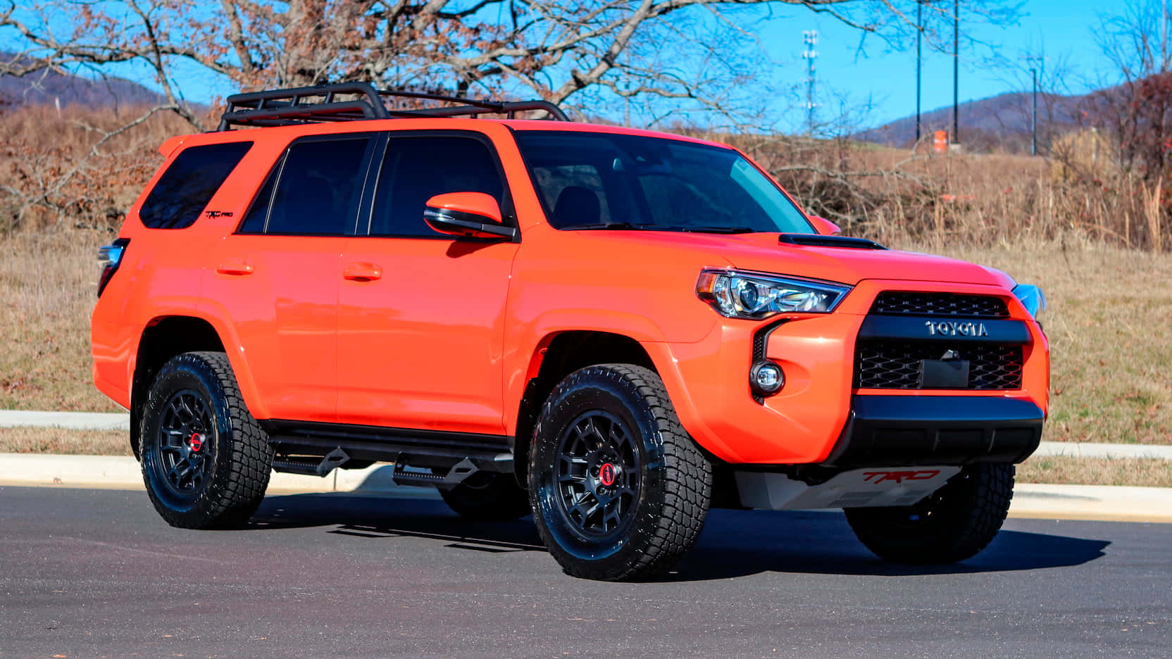 體驗2023款豐田4Runner的先進性能 壁紙 — 一輛充滿活力的橙色豐田 4Runner TRD 在柏油路上霸氣地停放，配備黑色輪圈和車頂行李架，背景是乾燥的草地山坡和晴朗的藍天。