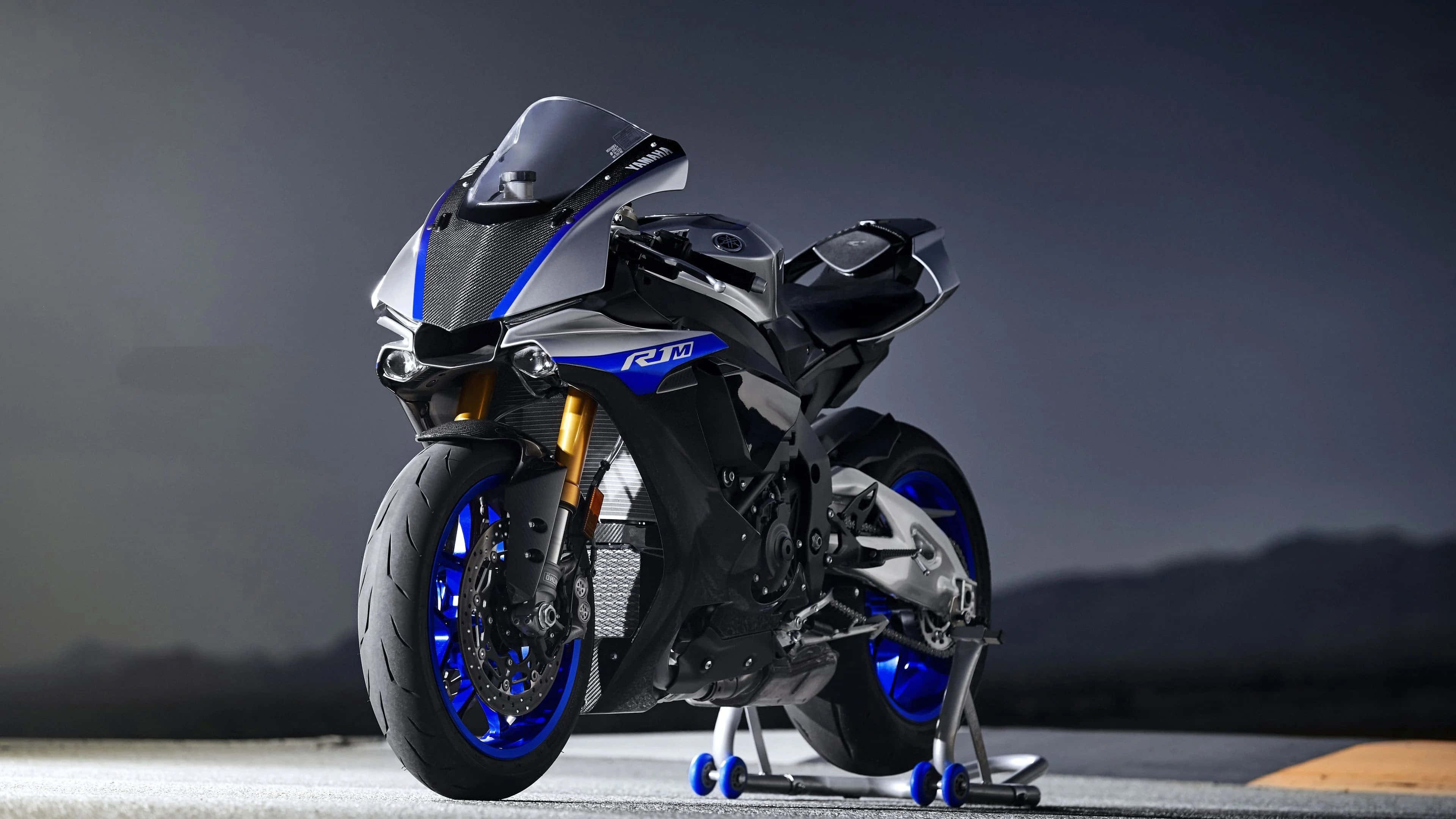 體驗4K自行車，暢遊公路 壁紙 — 一台時尚的Yamaha R1M運動型摩托車，帶有藍色點綴和金色前叉，在黑暗的山脈背景下蓄勢待發於賽道上。