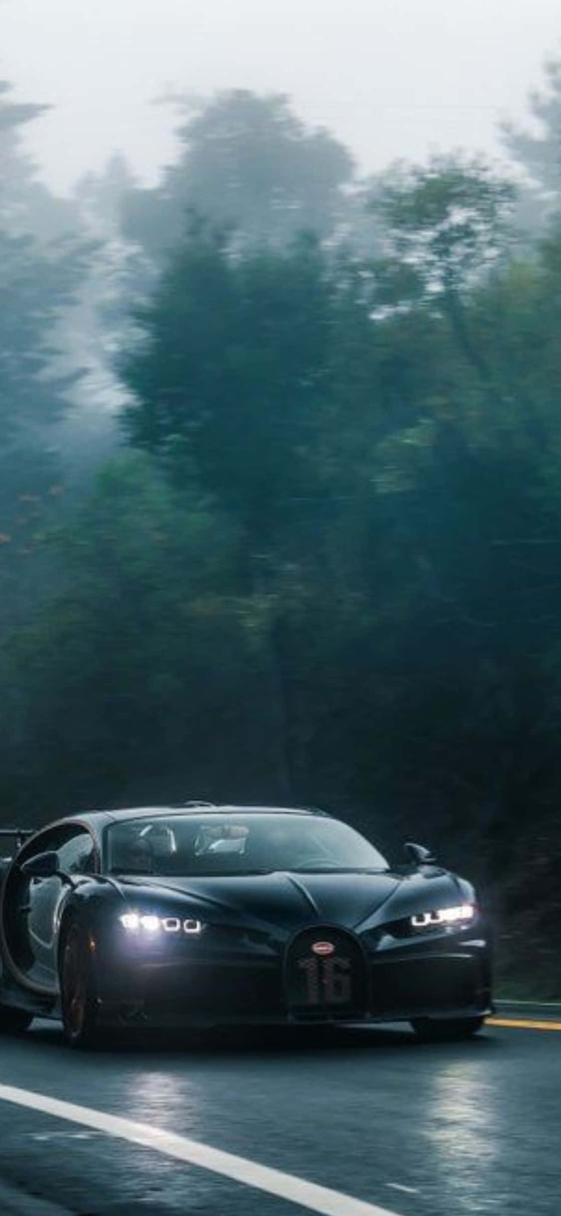 體驗iPhone XS與Bugatti的奢華 壁紙 — 一輛時尚的黑色布加迪Chiron在潮濕的森林道路上奔馳，周圍是霧氣籠罩的綠樹，車頭燈閃閃發光。