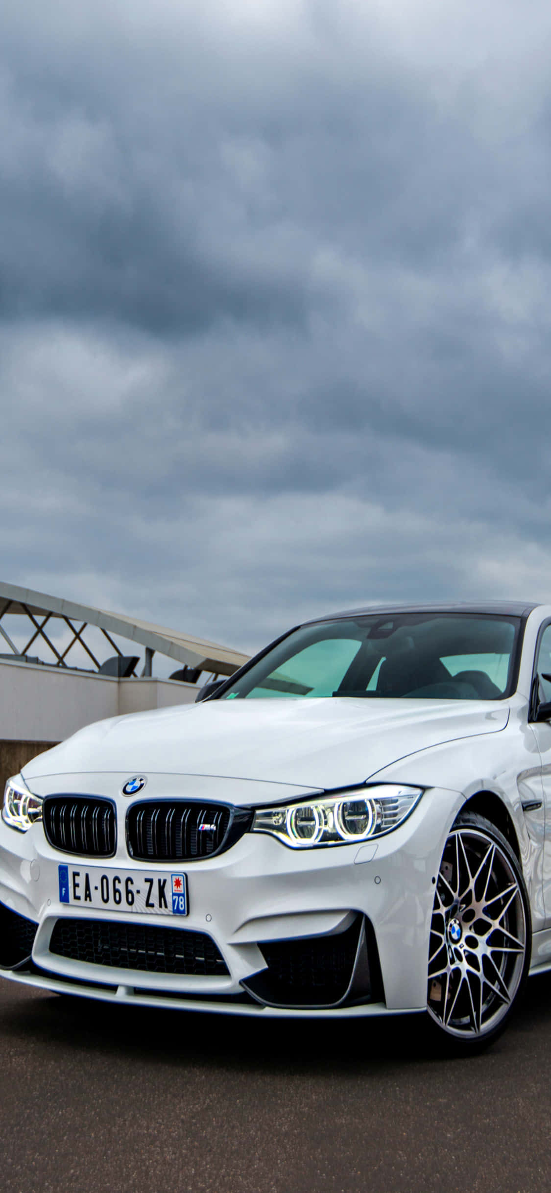 體驗iPhone X與BMW的強大性能 壁紙 — 一架時尚的白色BMW M4跑車停泊在戲劇性的多雲天空下，背景是現代建築。