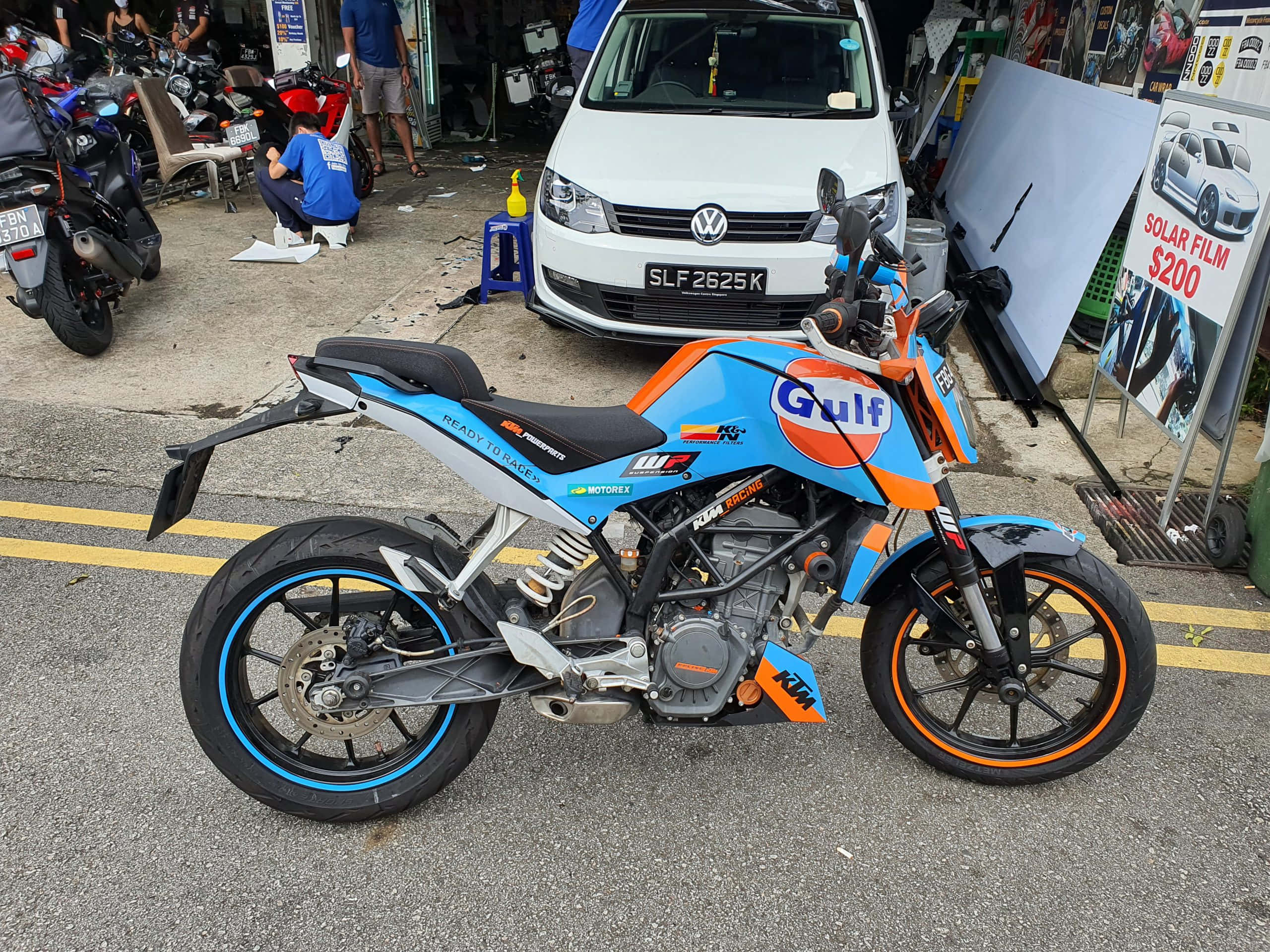 體驗KTM Duke的強勁動力——盡情釋放 壁紙 — 一架帶有Gulf塗裝的鮮豔藍色和橙色KTM電單車停泊在鋪砌的停車場上，旁邊有一輛白色客貨車和其他電單車。