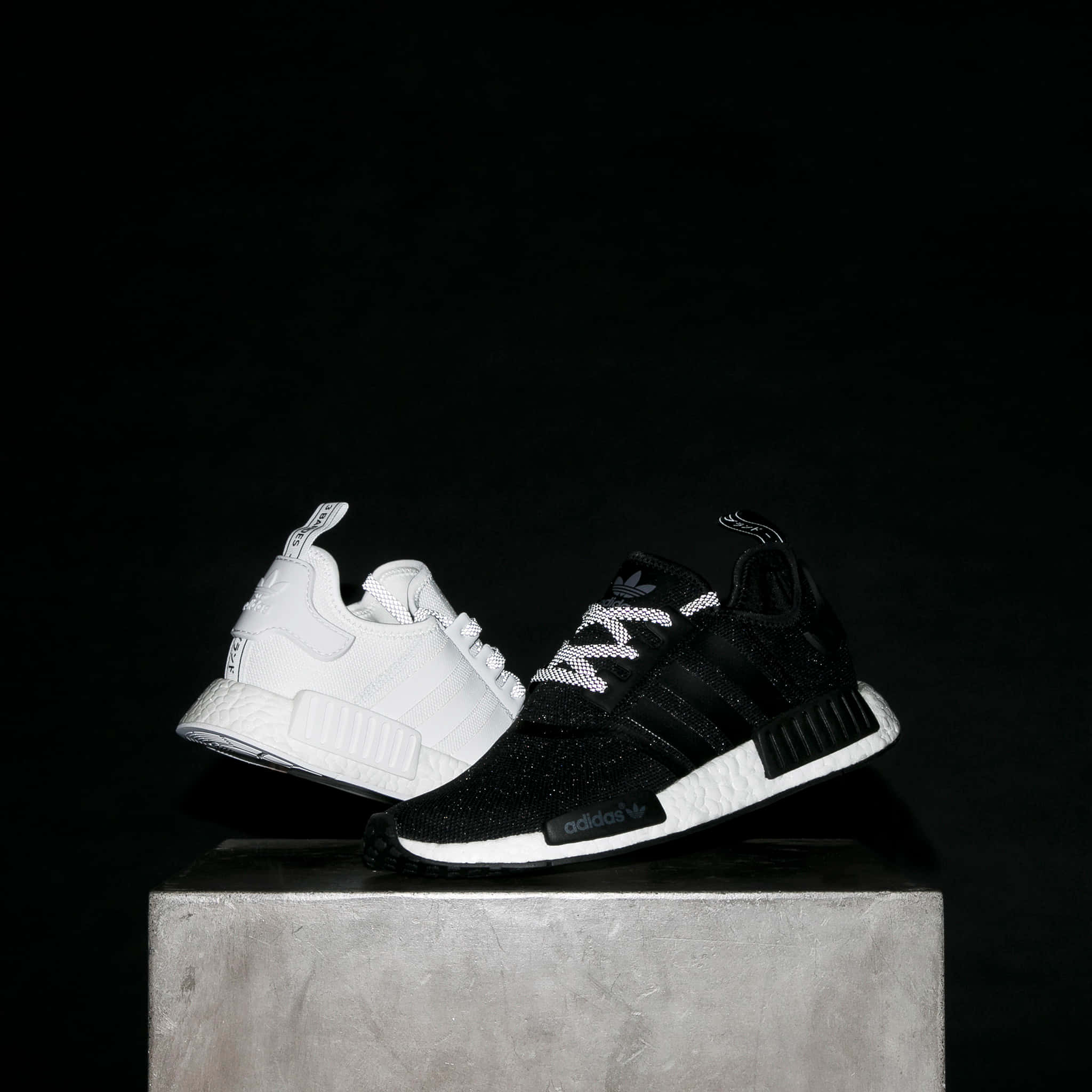 體驗Reebok的舒適與時尚 壁紙 — 兩對Adidas NMD運動鞋，一對白色一對黑色，放在灰色混凝土塊上，背景為純黑色。