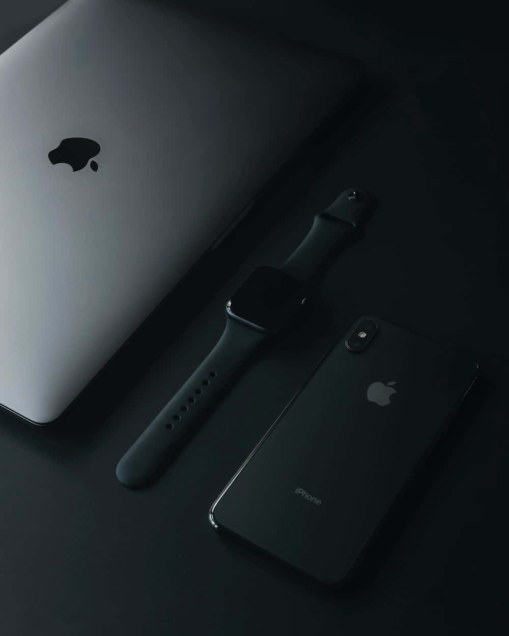 體驗全新 iPhone 的強大性能 壁紙 — 一部時尚的銀色 MacBook 手提電腦、黑色 Apple Watch 和黑色 iPhone 在柔和燈光下排列在深色表面上。