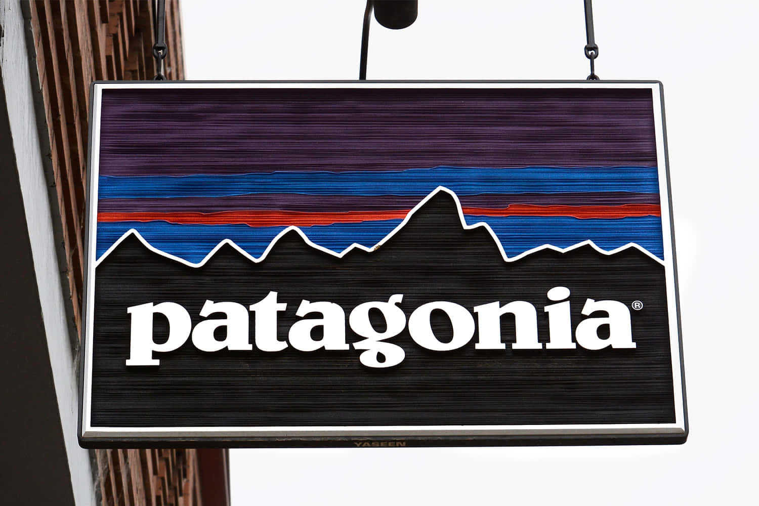 體驗巴塔哥尼亞之美——與自然連結的天堂 壁紙 — 一個懸掛的Patagonia商店招牌，特色是風格化的山脈，帶有紫色、藍色和紅色條紋，背景為黑色。