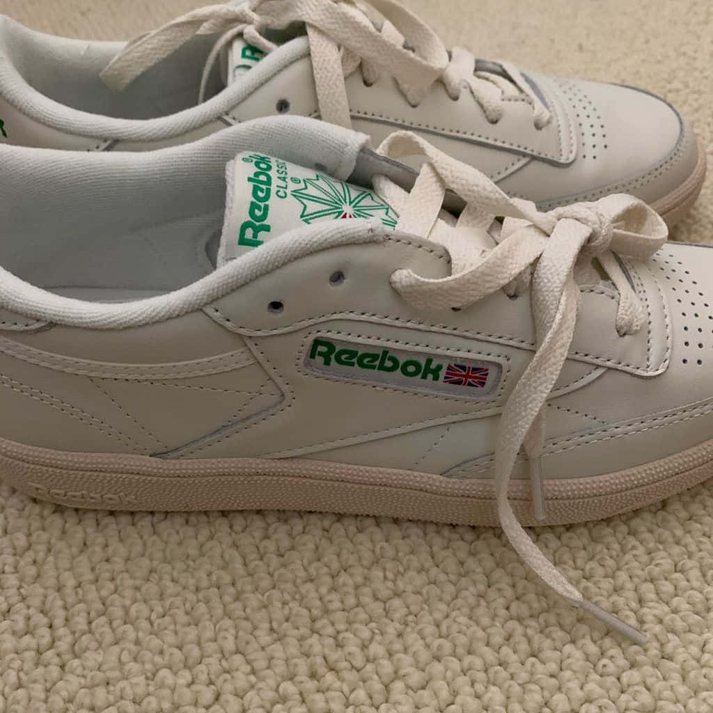 體驗彪馬經典風格 壁紙 — 一雙帶有綠色品牌標誌的白色 Reebok Classic 運動鞋特寫，放在有紋理的米色地毯上。