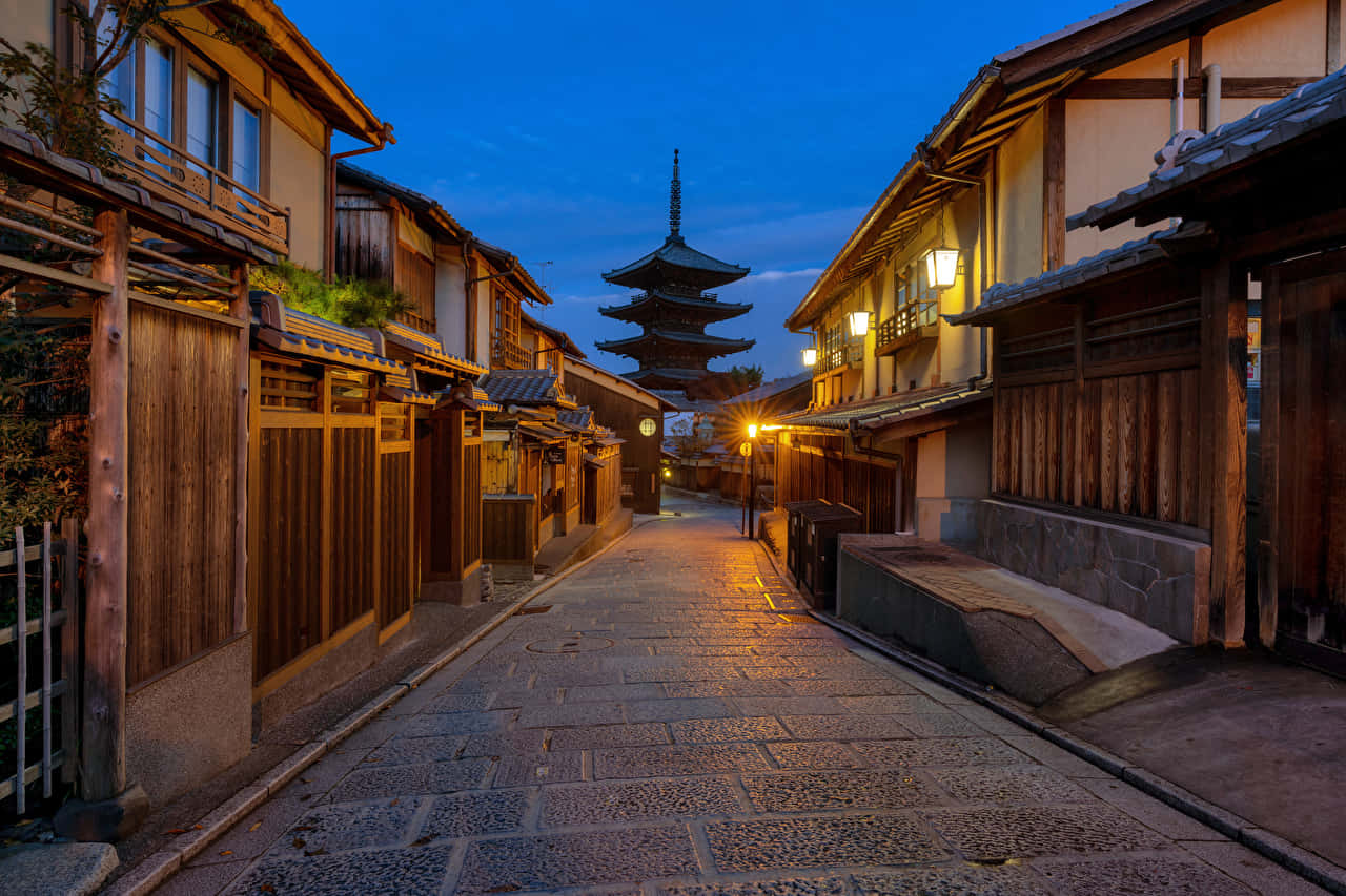體驗日本之美 壁紙 — 一條狹窄的石頭街道，兩旁是傳統木屋，通向暮色天空下發光的寶塔。